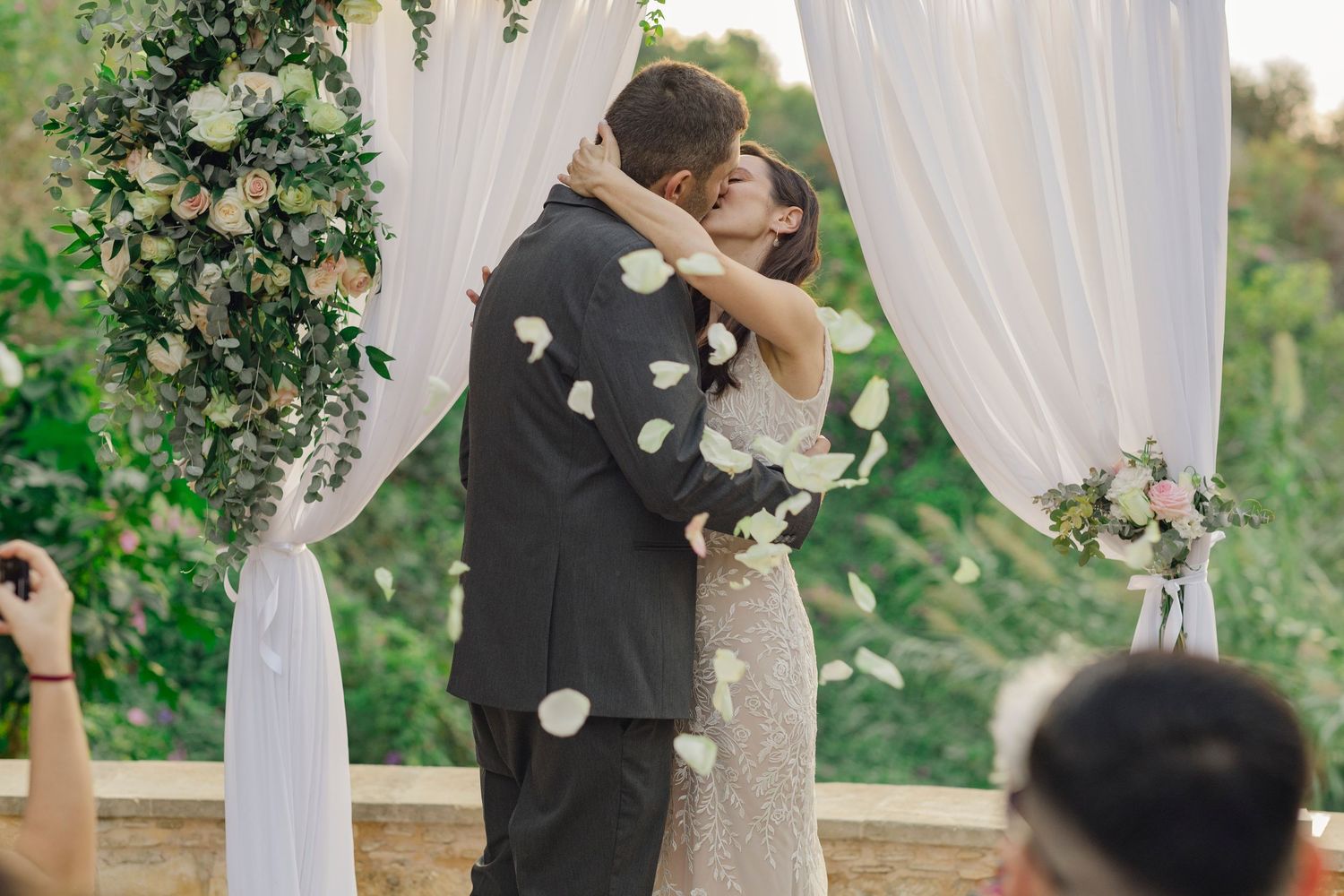 Couple sharing first kiss at outdoor wedding ceremony under floral arch with white draping and rose petals falling.