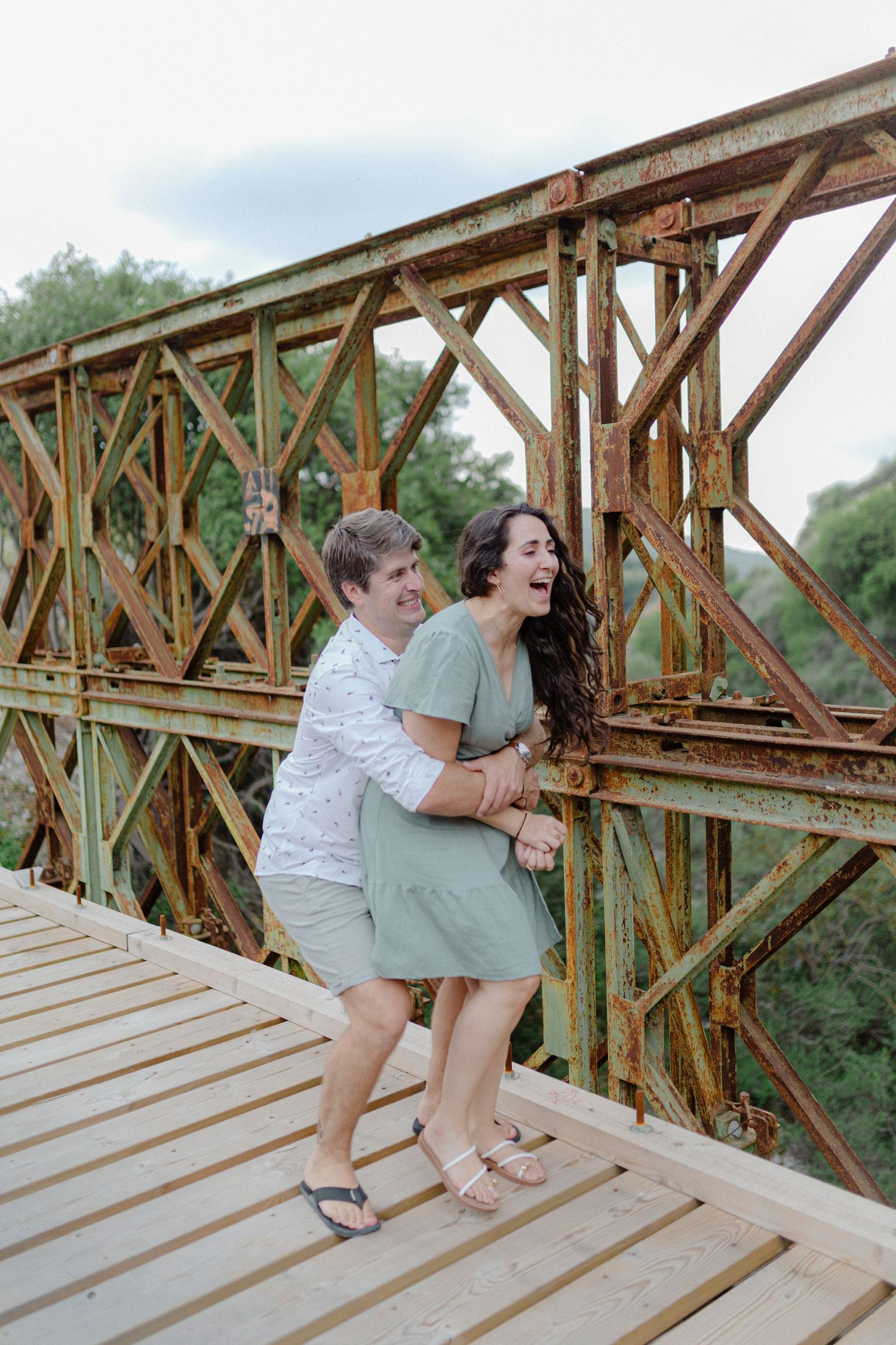 Couple laughing and embracing on a rustic wooden bridge with vintage iron trusses outdoors.