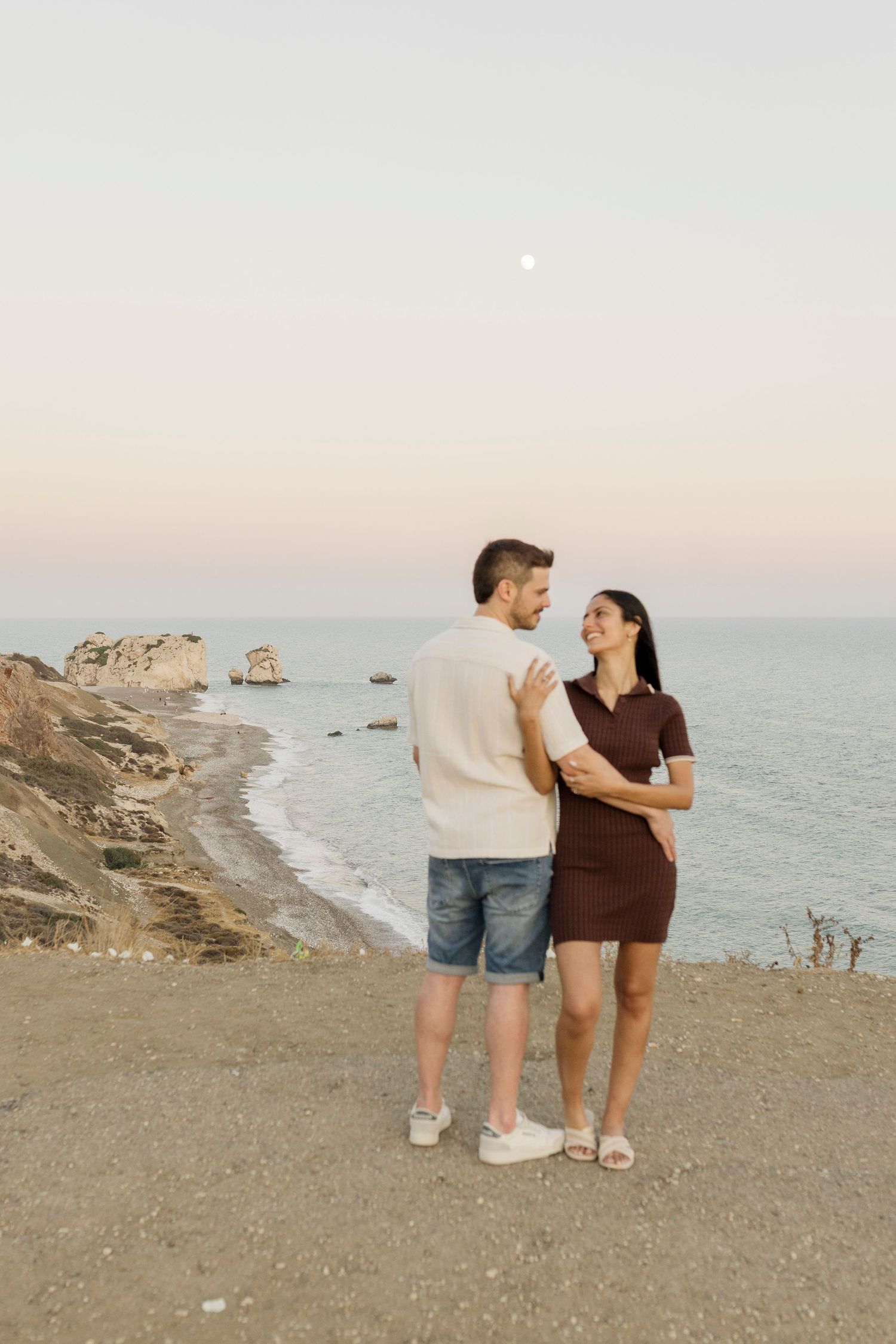 Couple embracing on coastal cliff at sunset, woman in brown dress, man in white shirt and denim shorts, ocean backdrop.