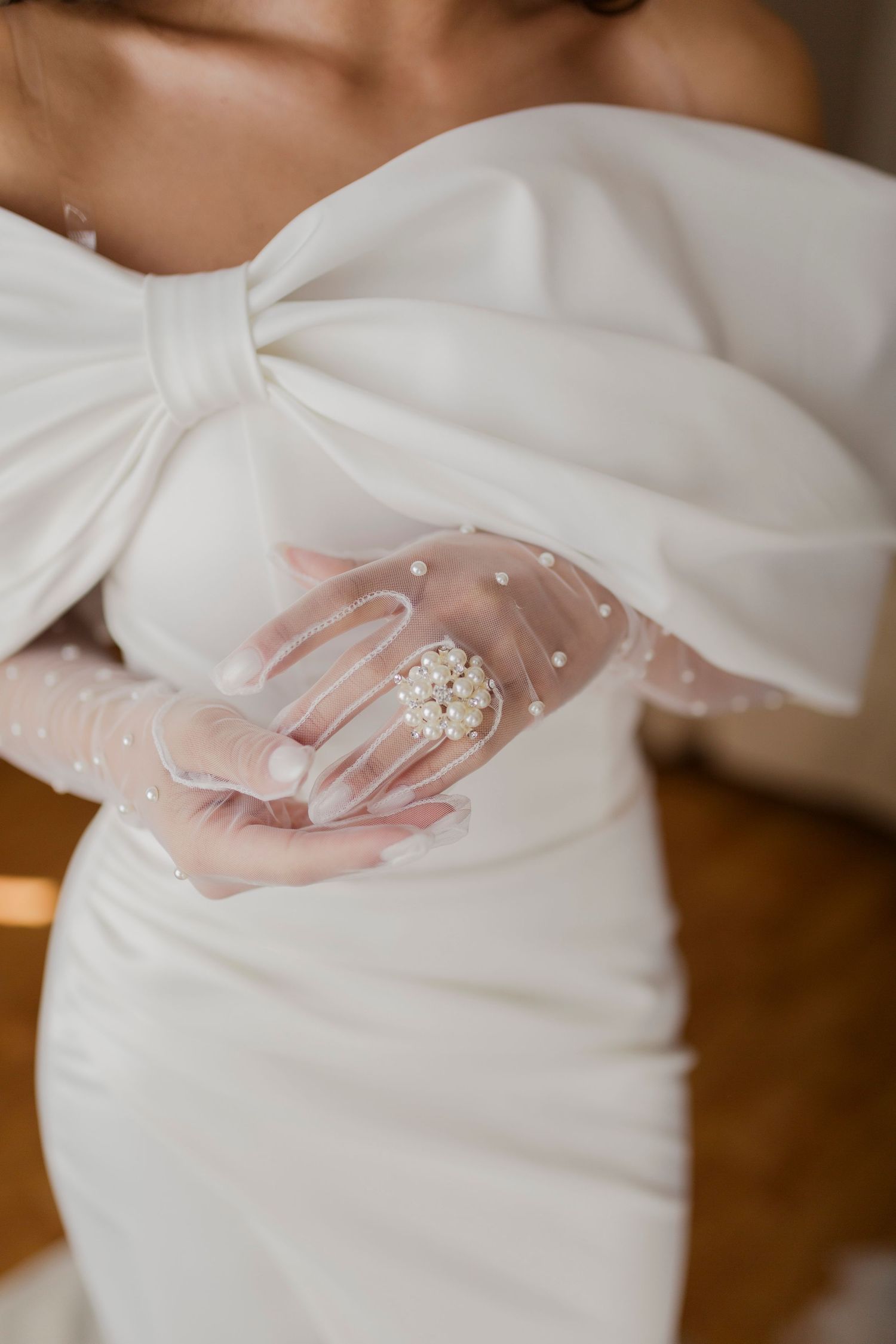 Bride in white off-shoulder gown with large bow detail wearing sheer pearl-embellished gloves holding a floral brooch.