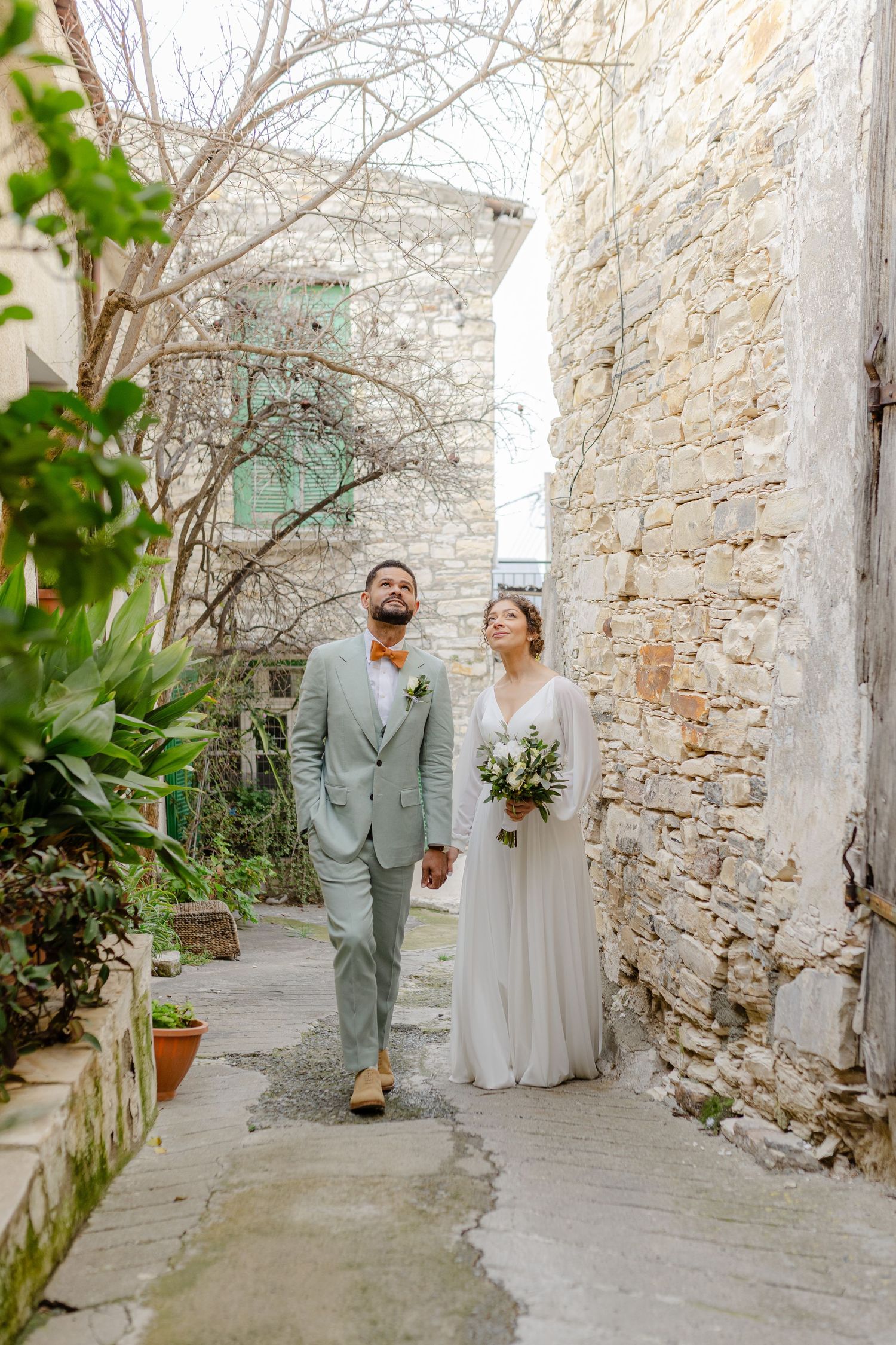 Bride and groom walk hand-in-hand through a charming stone alley, she in white gown, he in gray suit with orange bow tie.