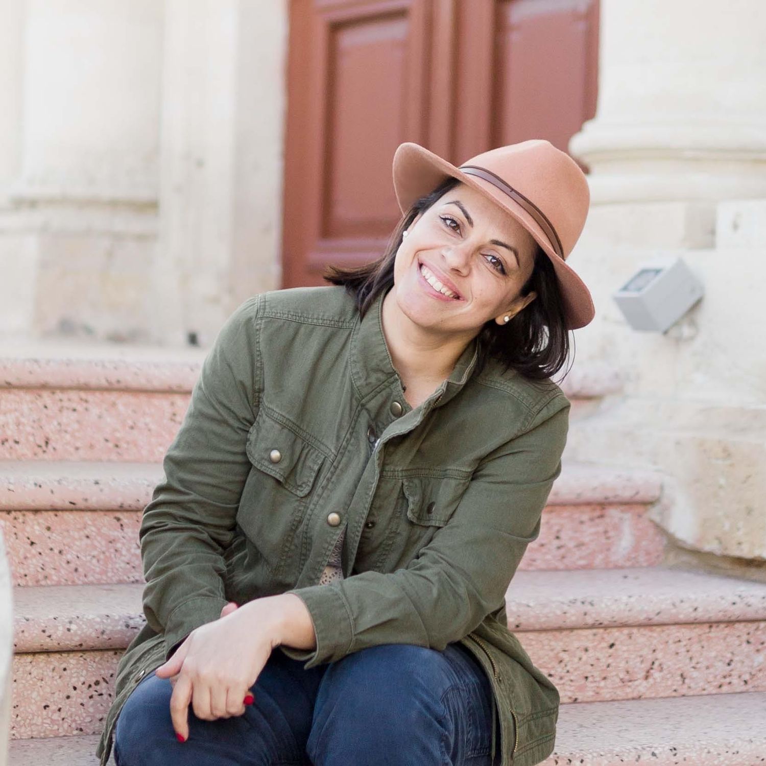 Smiling woman in green jacket and tan hat sitting on stone steps in an outdoor setting.