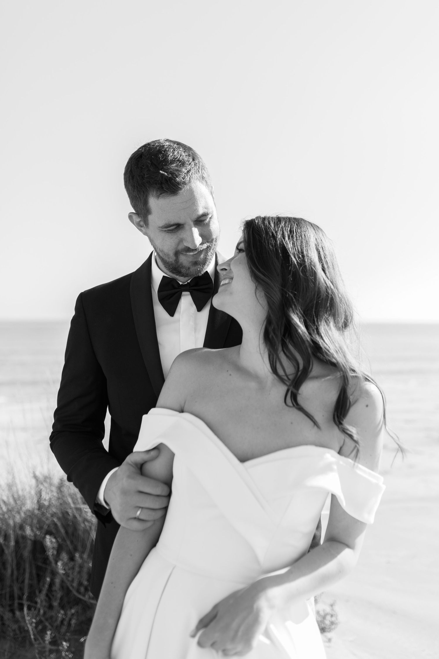 Black and white romantic portrait of bride and groom embracing near ocean shoreline.