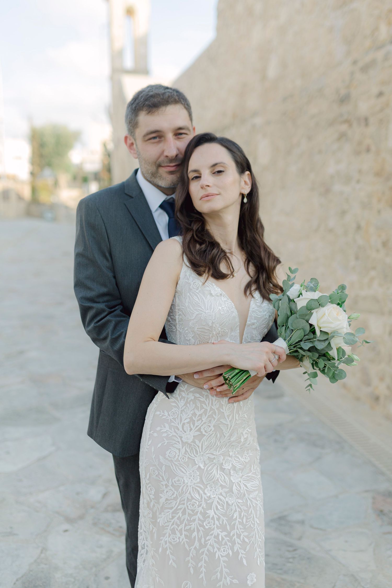 A bride and groom pose together outdoors, she holds a white rose bouquet in a lace gown as he embraces her in a gray suit.
