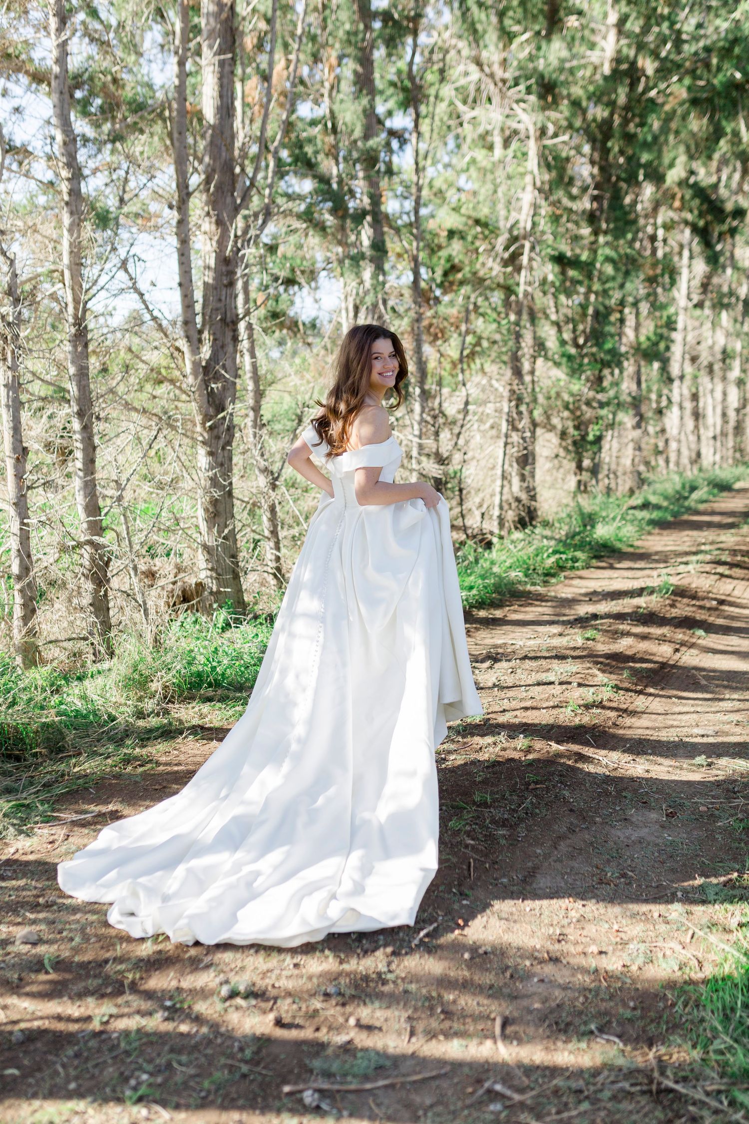 Elegant bride in off-shoulder white gown with long train posing on a sunlit forest path.