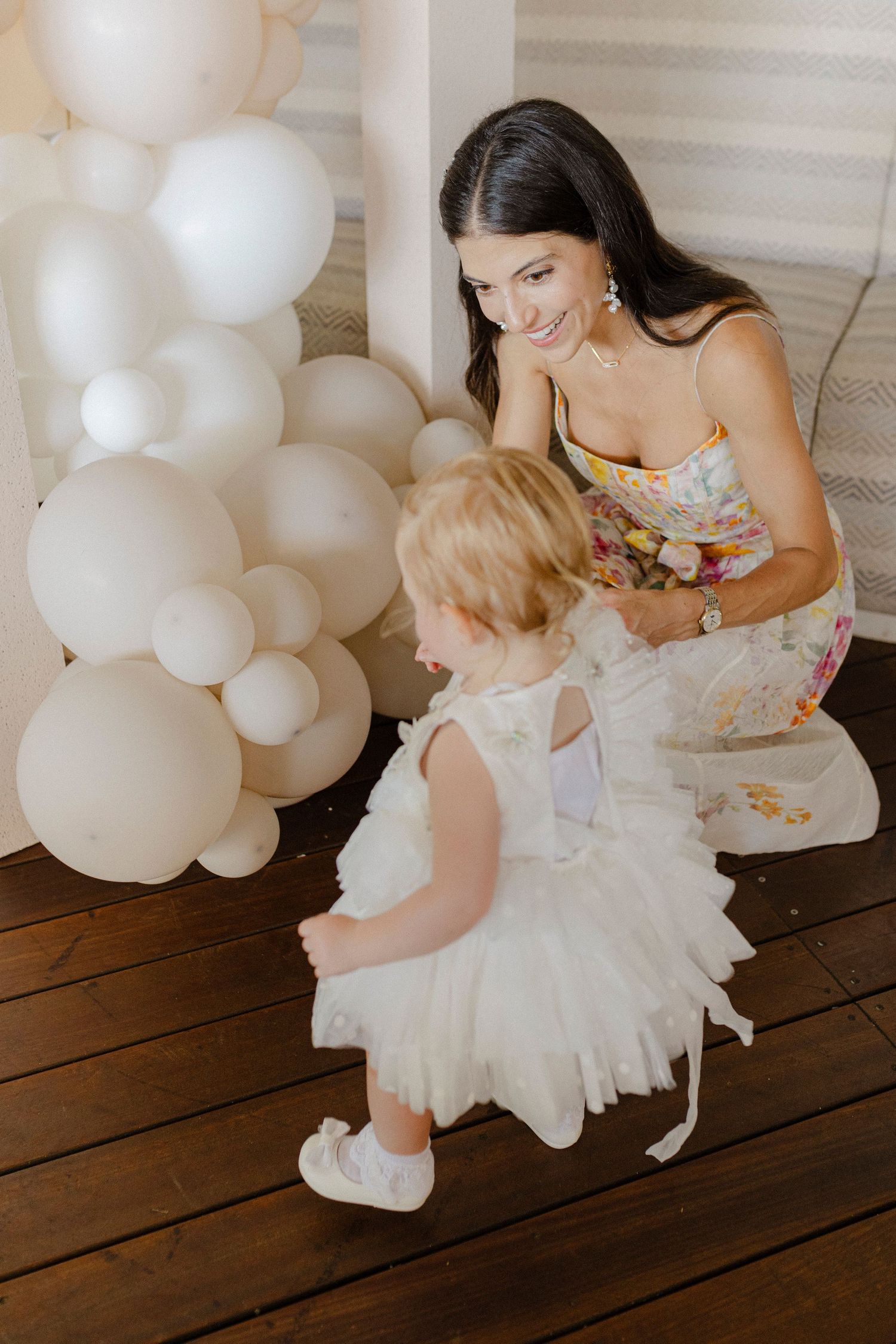 Smiling dark-haired woman in floral dress crouches near white balloons with toddler girl in white tutu dress.