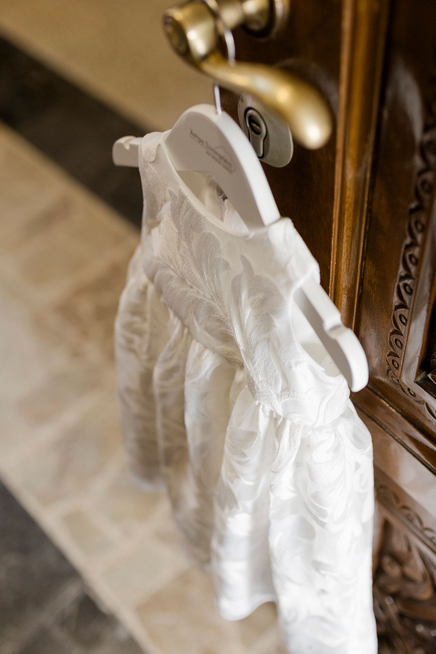 White lace flower girl dress hanging on ornate wooden cabinet door with gold hardware.