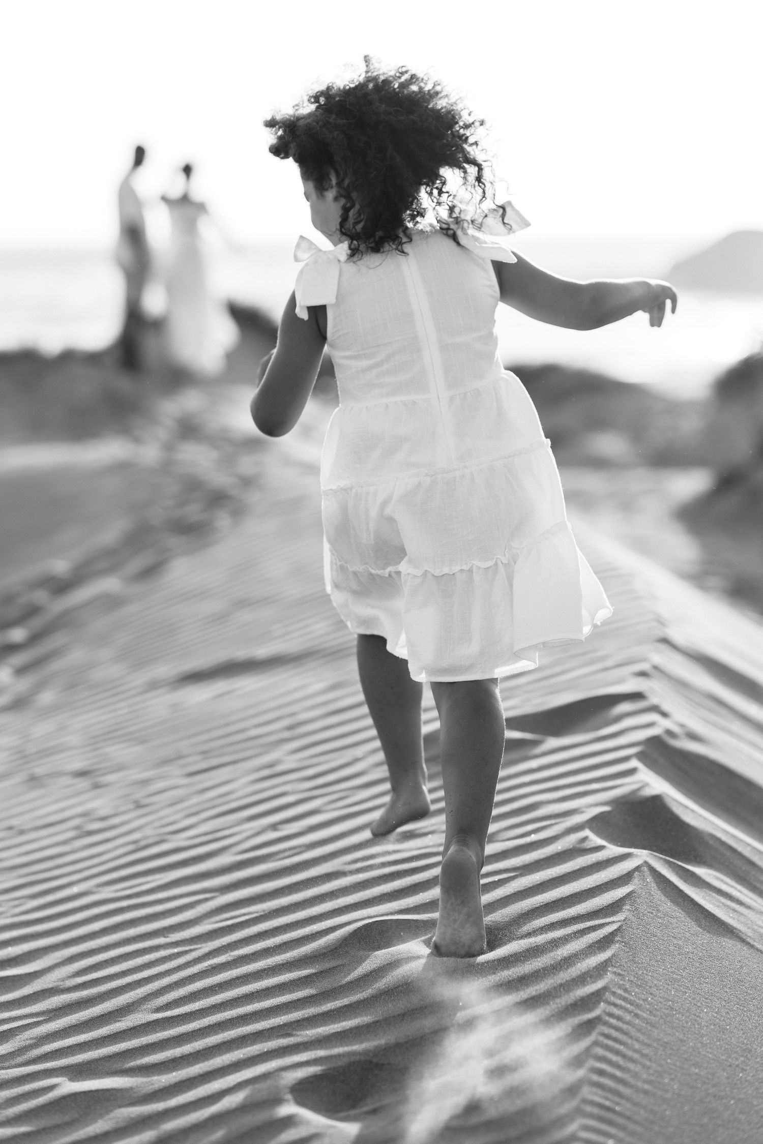 Black and white photo of a young girl in a white dress dancing barefoot on sand dunes near the beach.