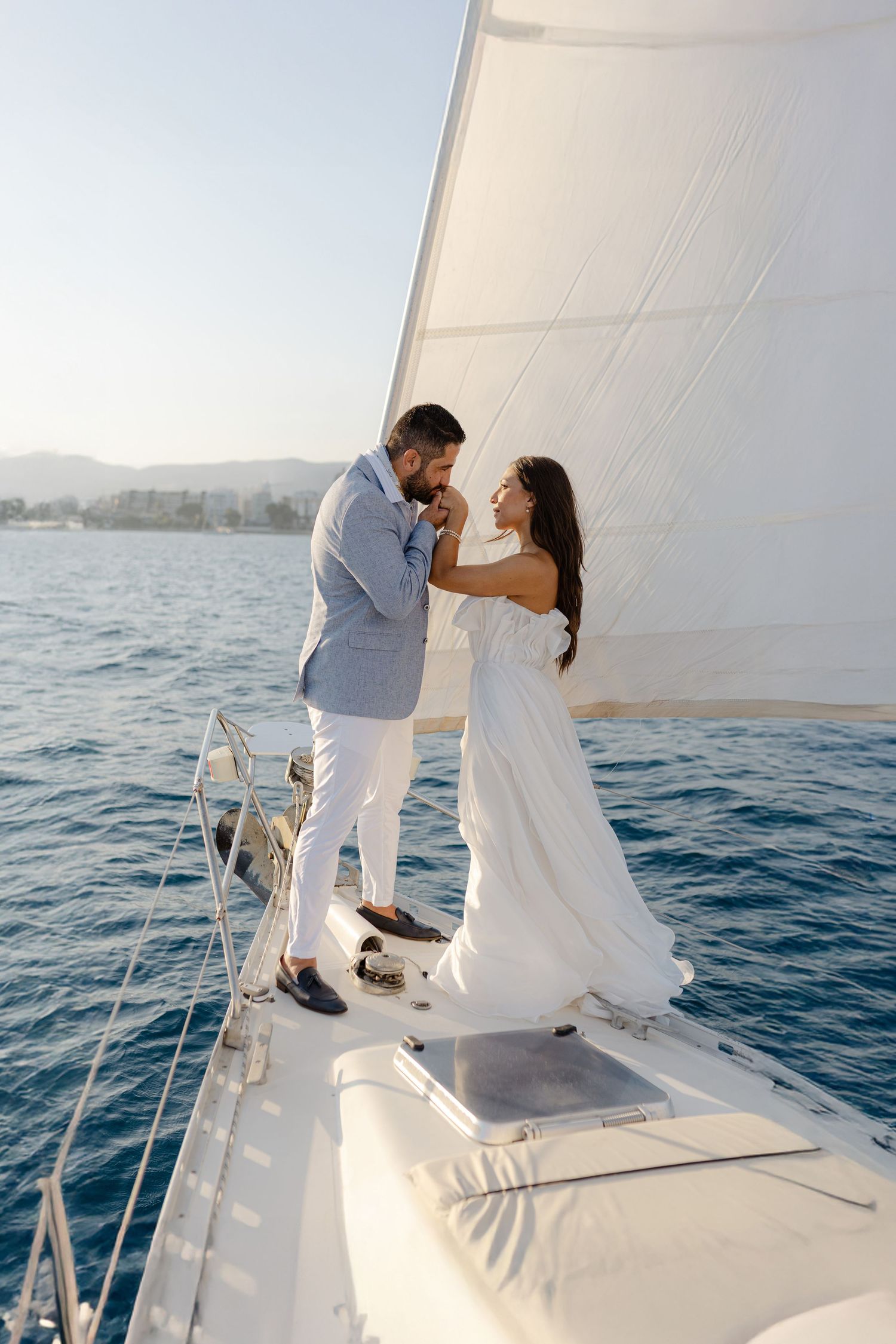 Romantic couple in elegant attire standing on a white sailboat deck with ocean views on a sunny day.