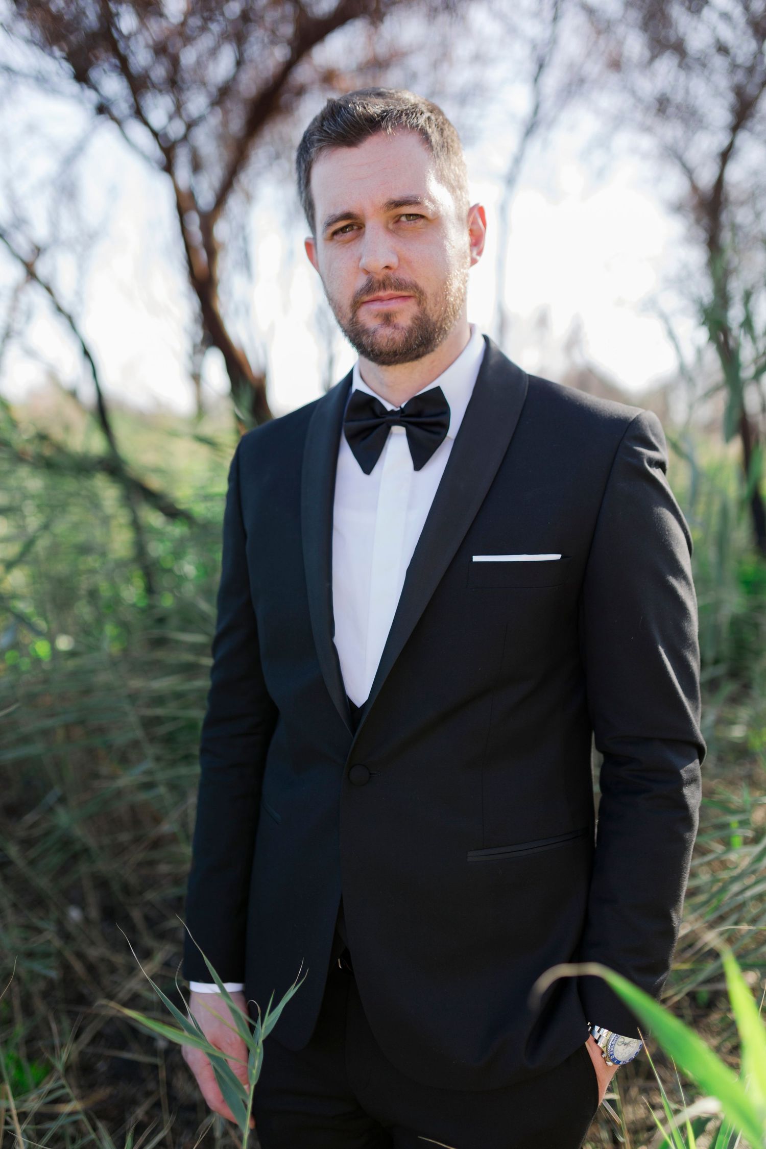 Elegant groom in black tuxedo with bow tie and pocket square posing outdoors among green foliage.