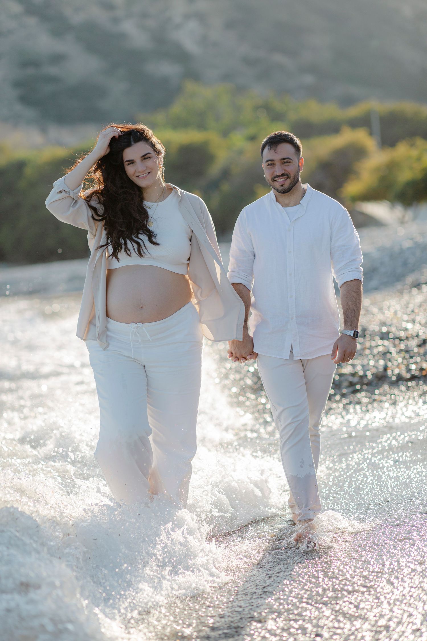 Pregnant couple in white outfits walking hand in hand along a sunny beach shoreline with waves splashing around their feet.