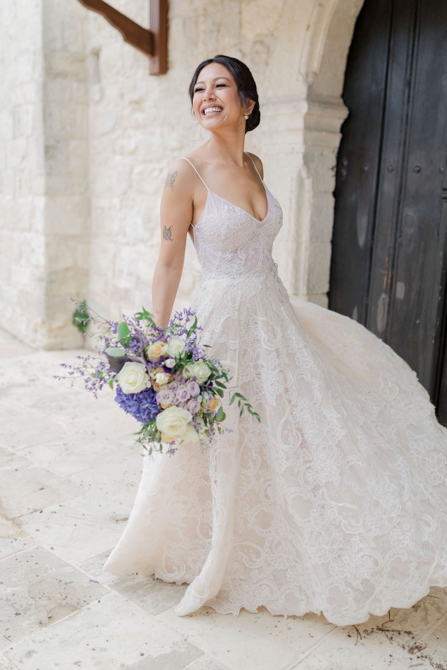 Joyful bride in lace ballgown with purple and white bouquet laughing outside stone building with black door.