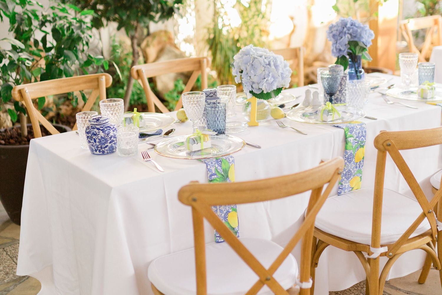 Elegant wedding table setting with blue hydrangeas, blue and white vases, lemon napkins, and wooden cross-back chairs.