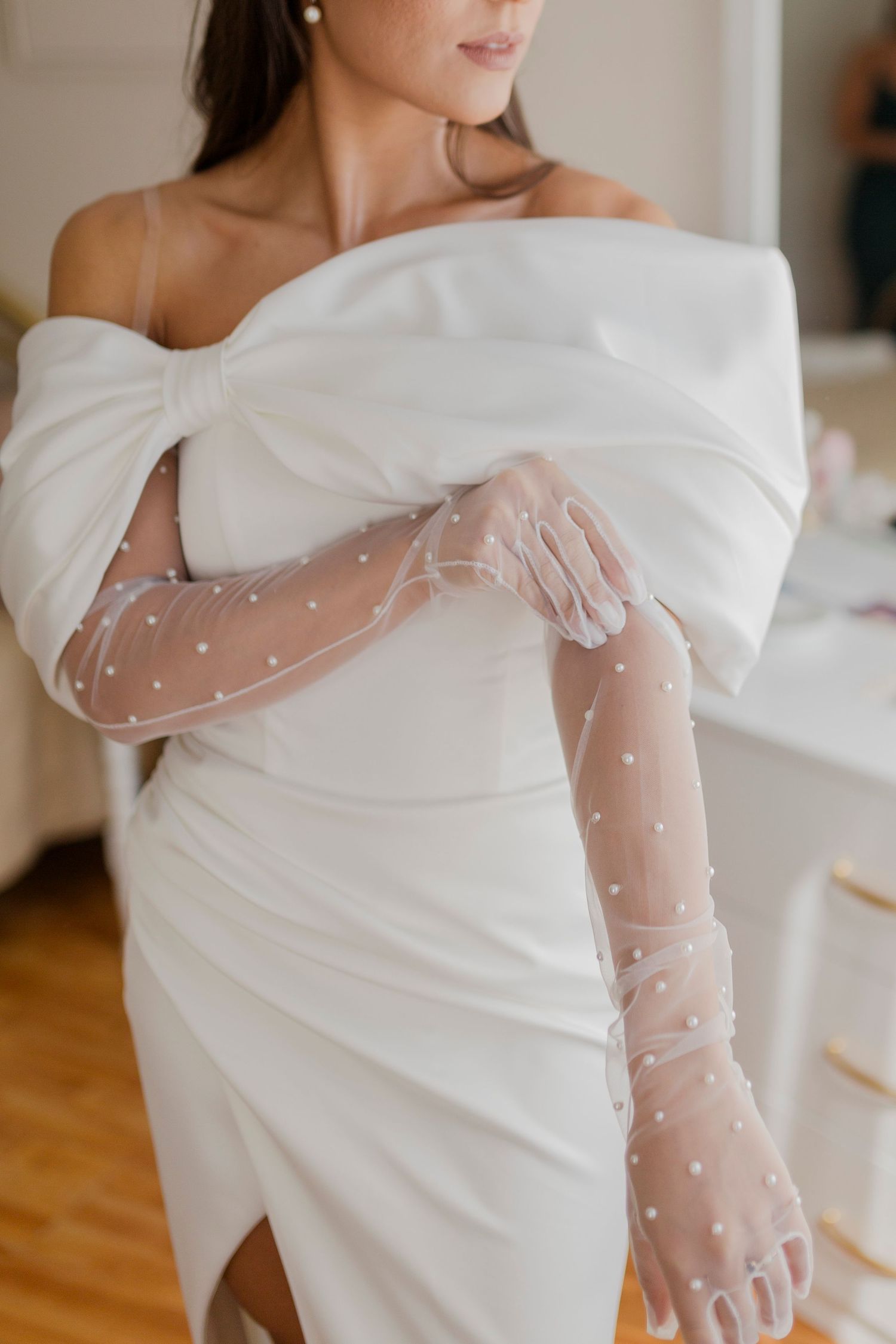 Bride wearing a white off-shoulder gown with a bow and sheer pearl-embellished long gloves.