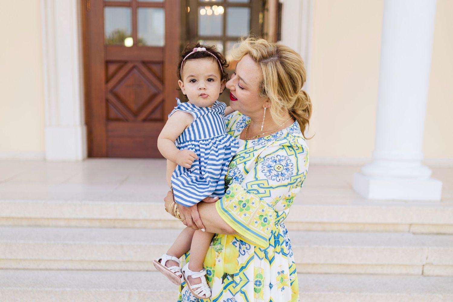 Woman in floral dress holding a baby girl in blue striped outfit outside elegant building.