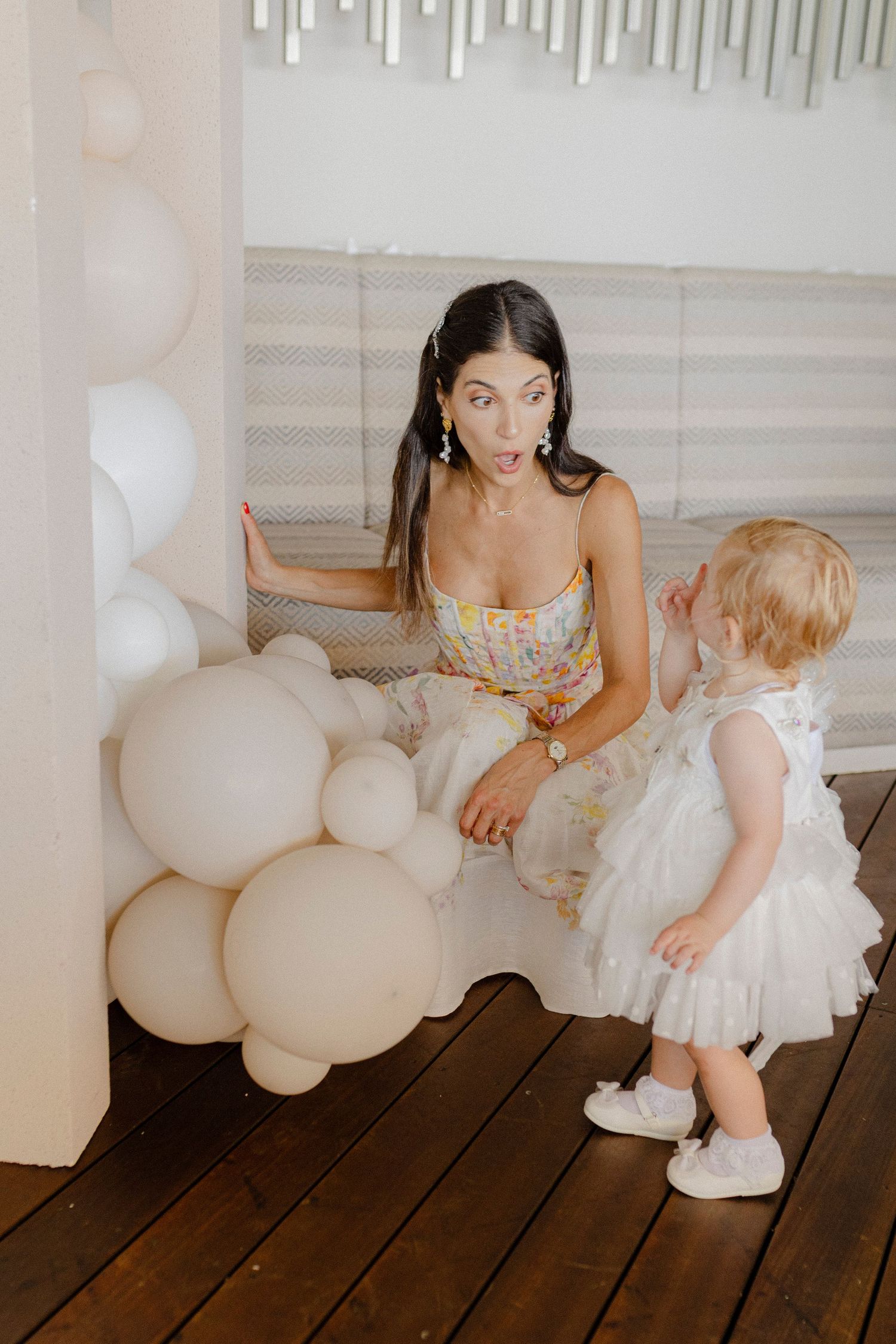 A woman in a floral dress reacts with surprise to a toddler in white dress surrounded by cream balloons indoors.