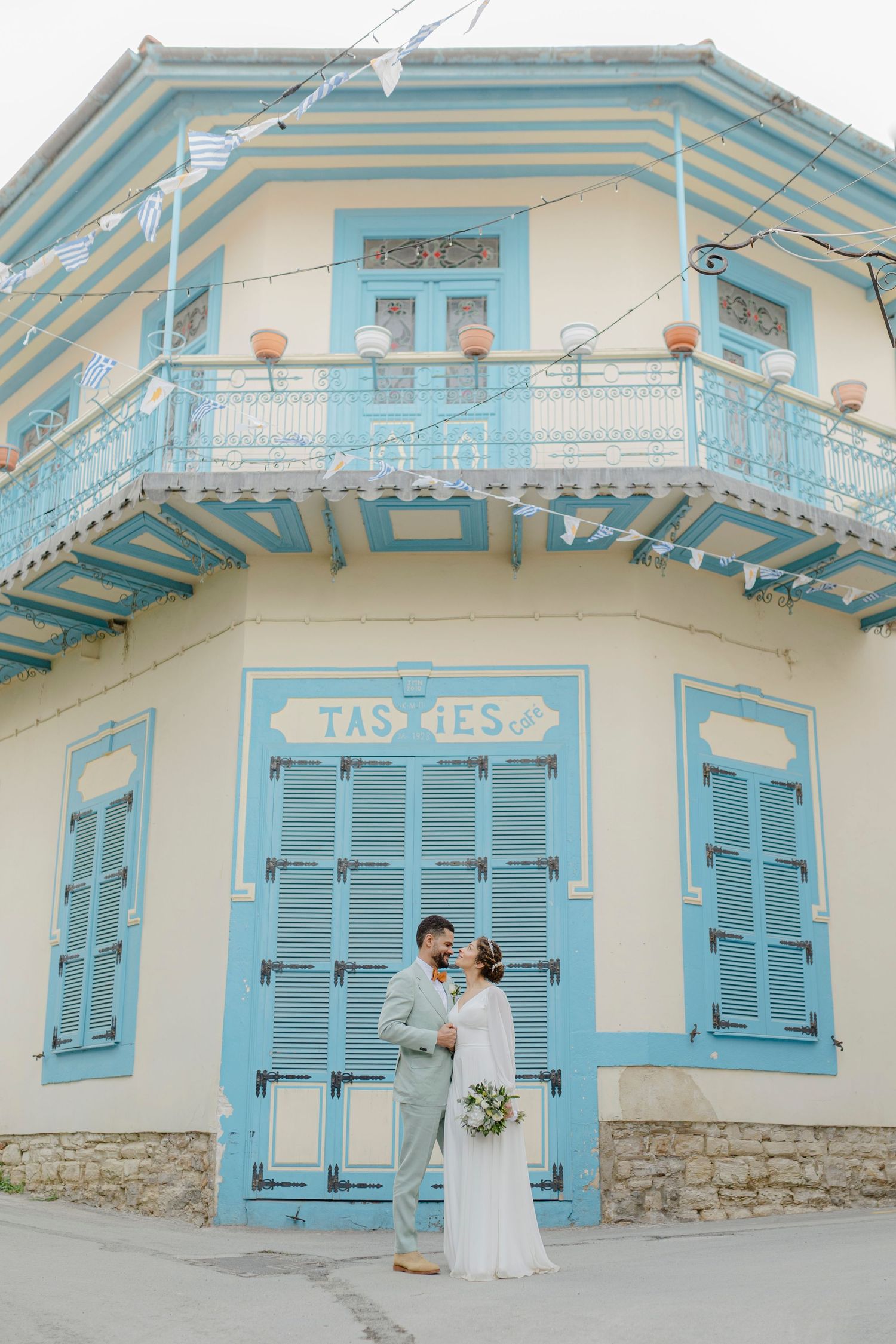 Wedding couple poses before colorful blue and yellow Greek building with shuttered windows.
