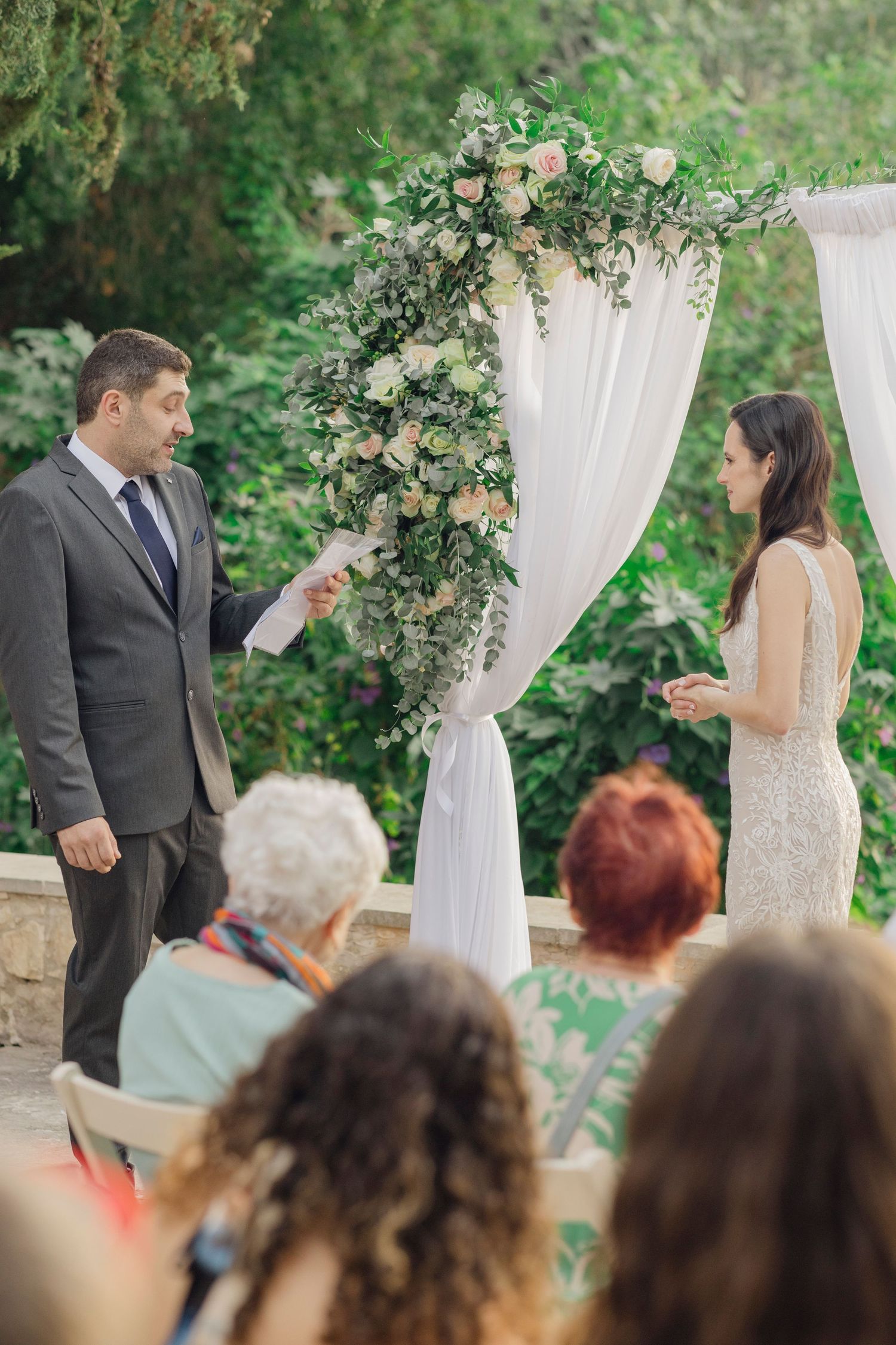 Outdoor wedding ceremony with couple exchanging vows under floral arch with white draping and seated guests.