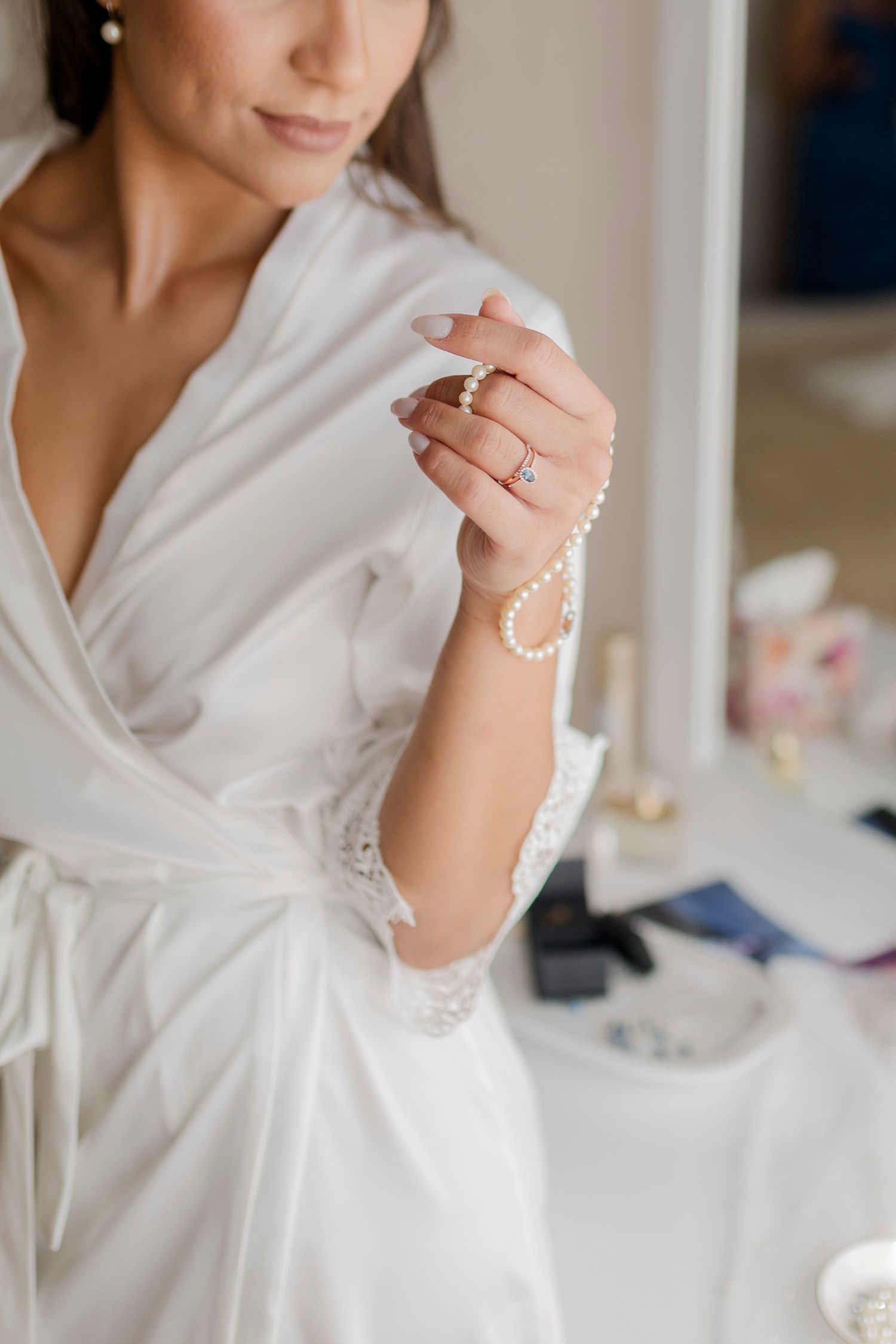Bride in white lace robe holding a delicate pearl bracelet while sitting at a vanity mirror.