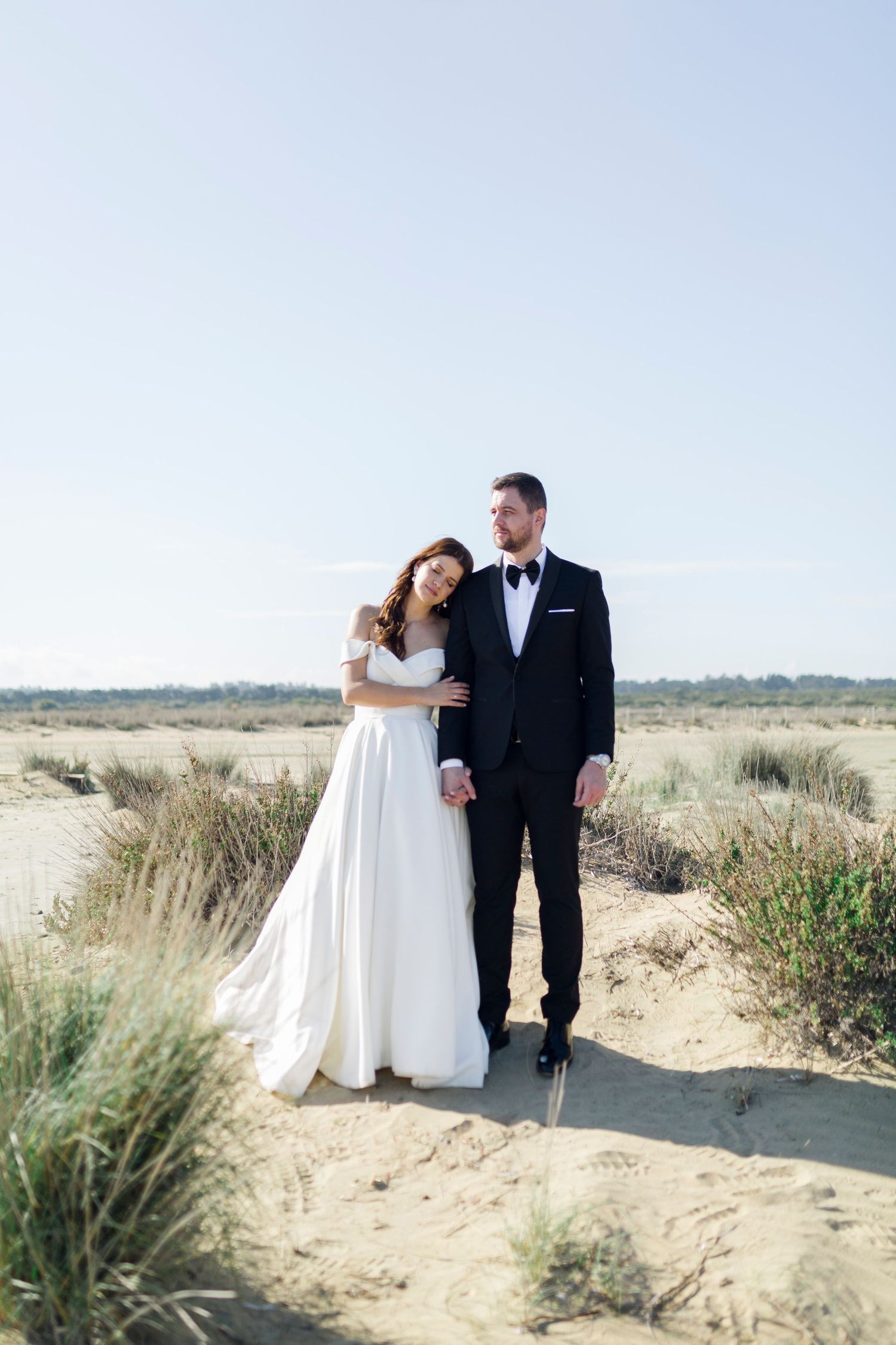 Bride in white off-shoulder gown and groom in black tuxedo pose together on sandy beach dunes.