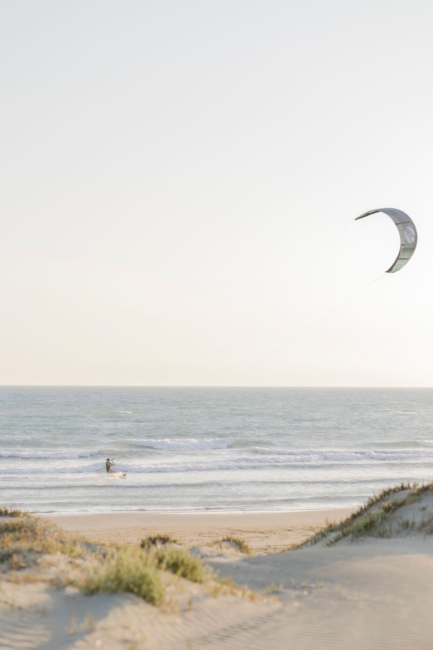 Kitesurfer rides ocean waves on a sandy beach at golden hour with kite soaring in pale sky.