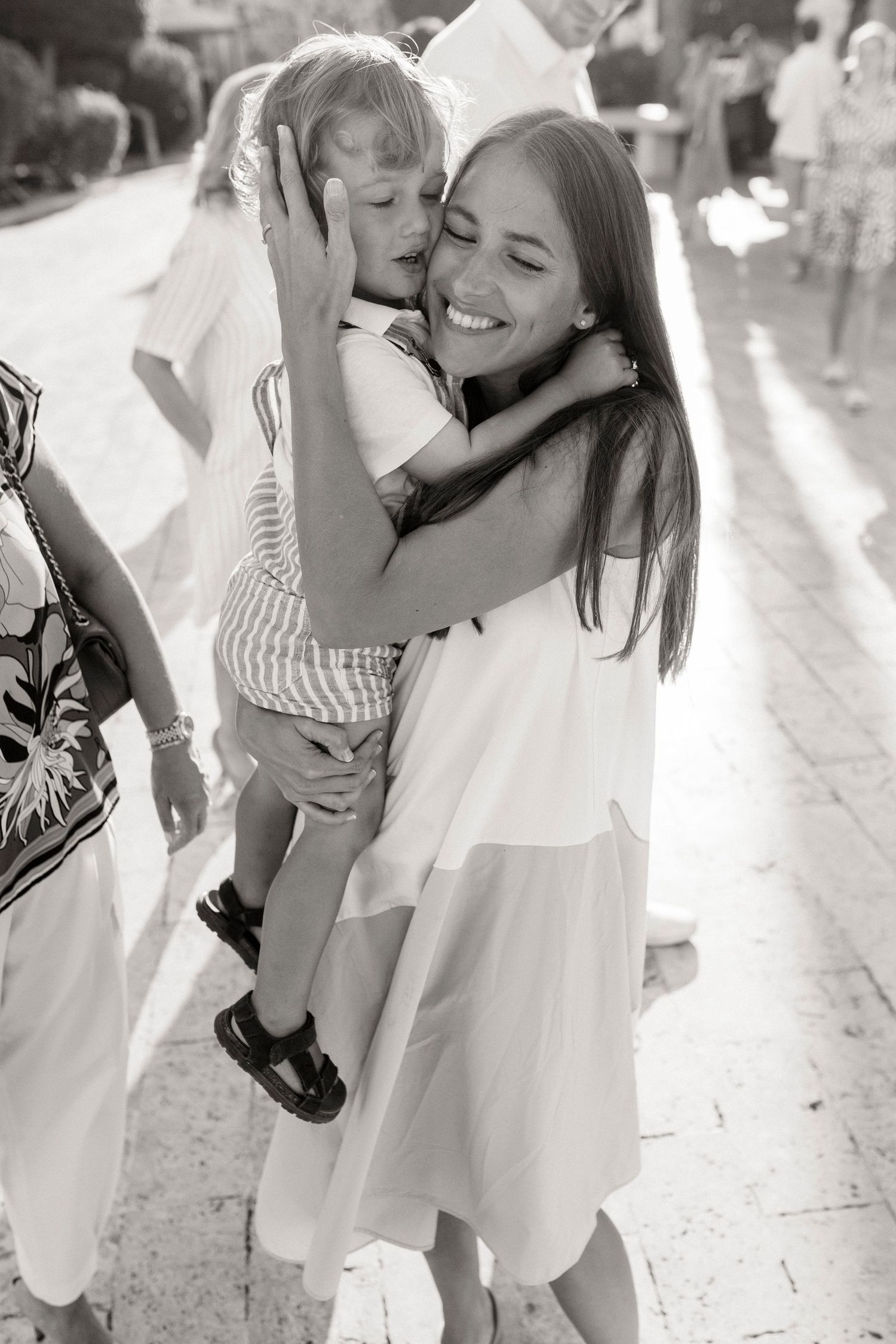Black and white photo of a smiling woman in a white dress hugging a young child outdoors.