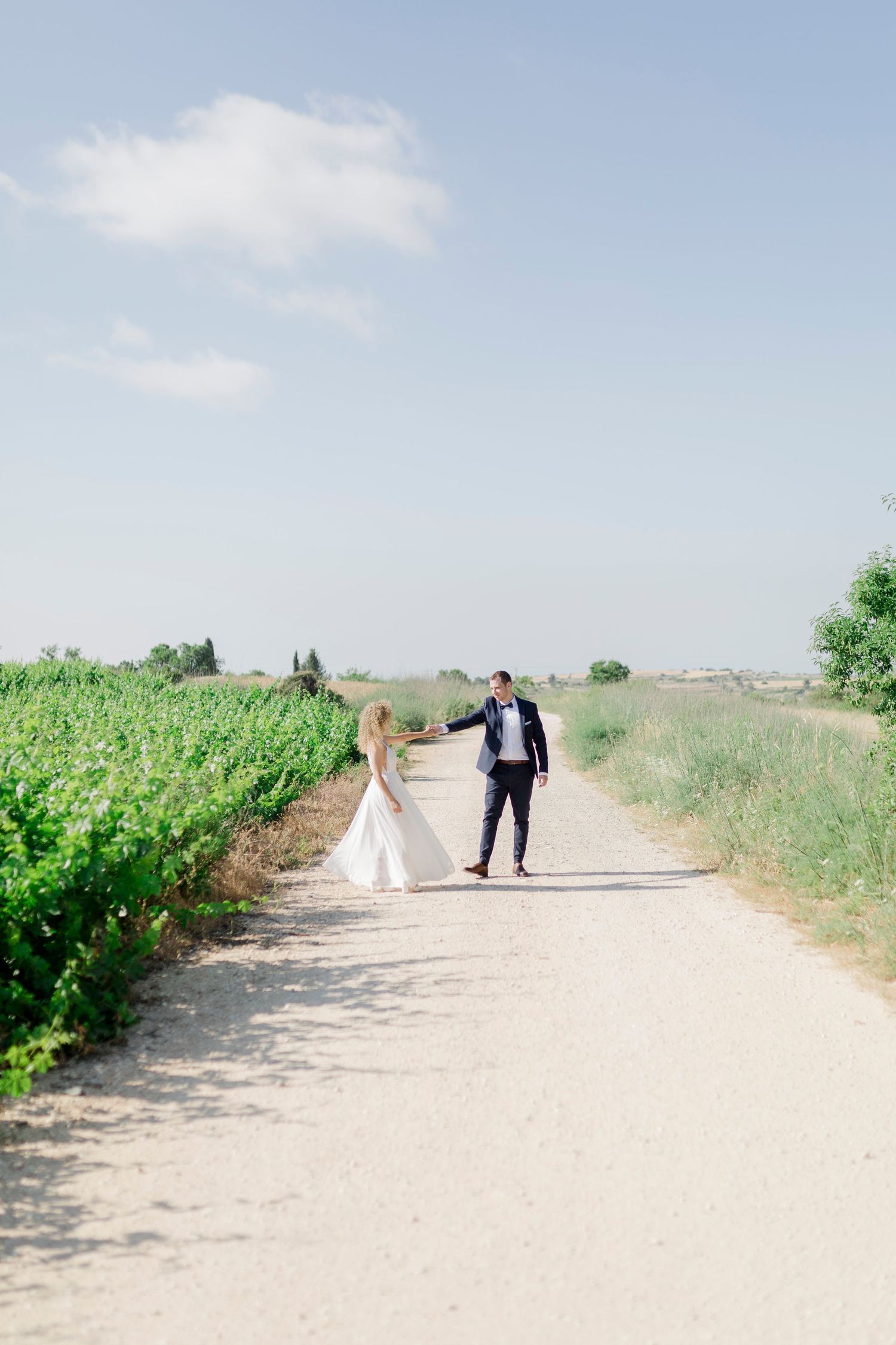 Bride and groom holding hands walking down a sunny countryside path lined with green fields on their wedding day.