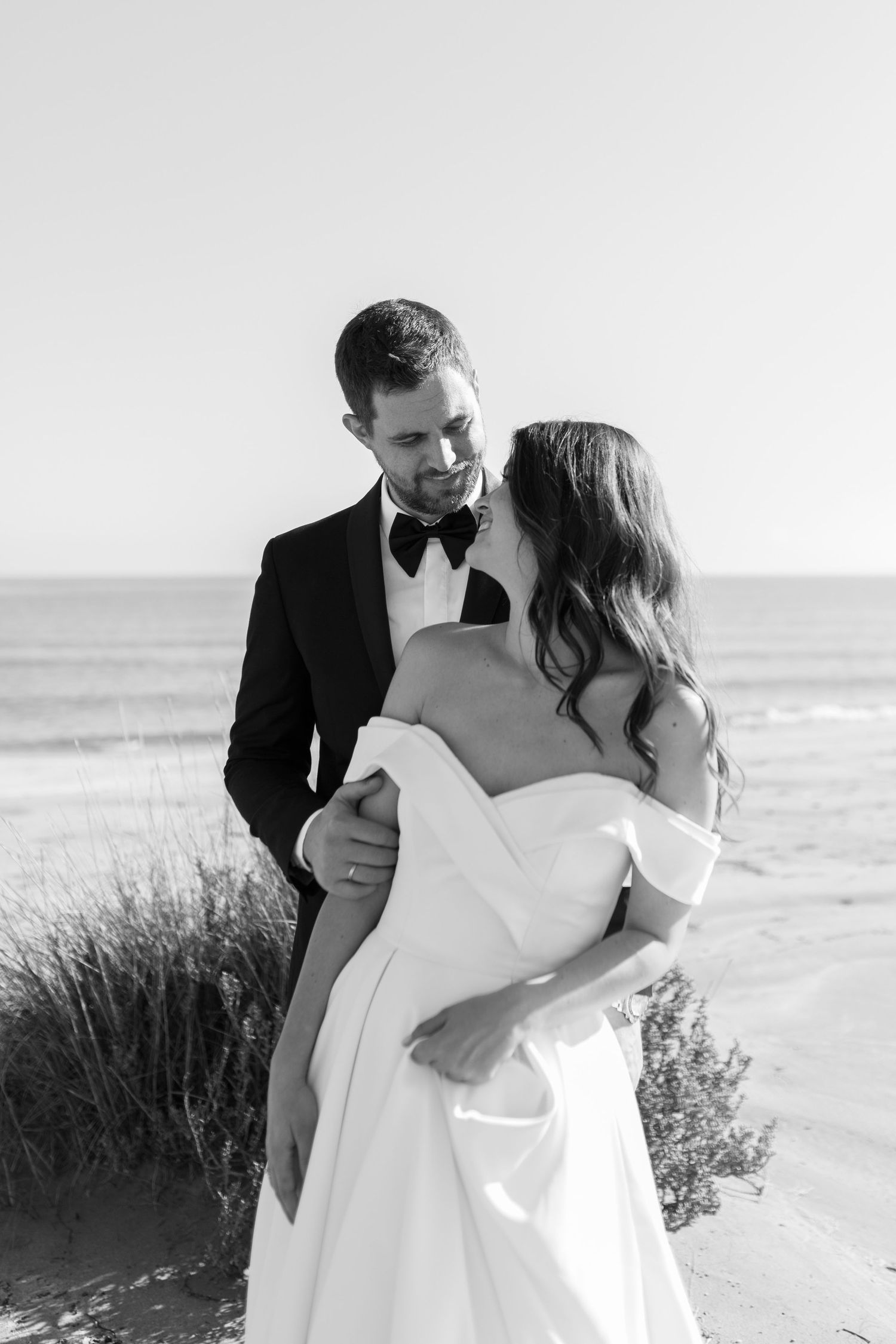 Black and white beach wedding photo of bride and groom embracing near ocean dunes, groom in tuxedo, bride in off-shoulder gown.
