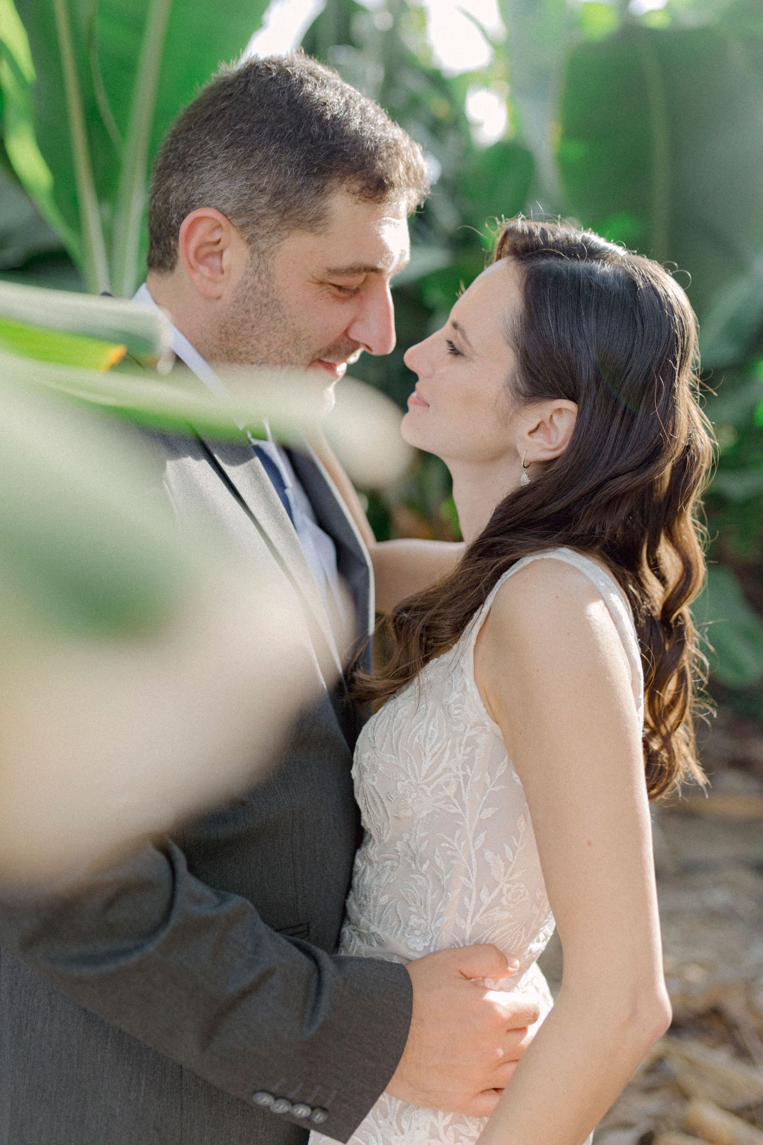 A bride and groom share a romantic moment, nearly kissing, surrounded by lush tropical greenery.
