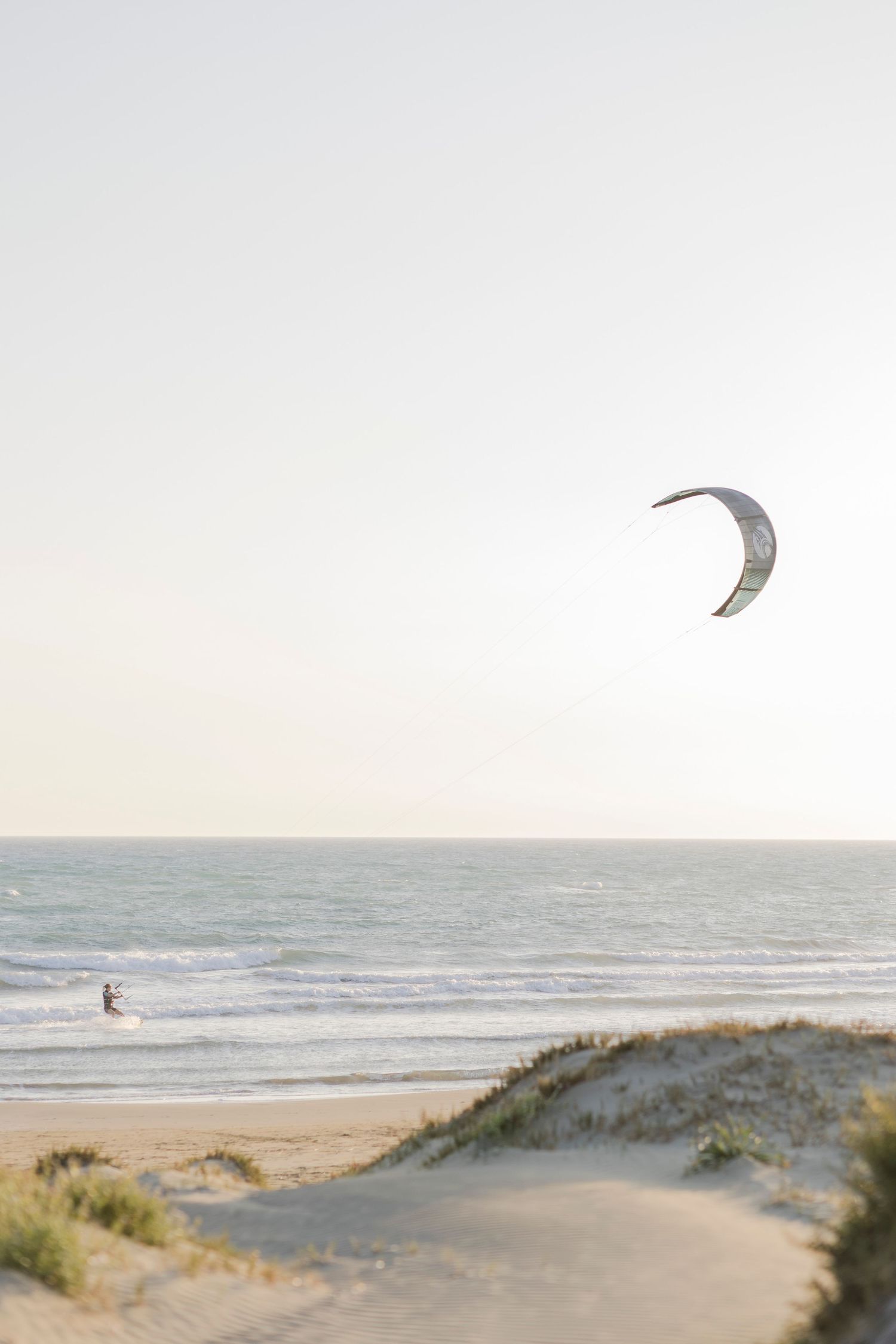 A kite surfer rides ocean waves near sandy dunes under a pale sky at a serene beach.