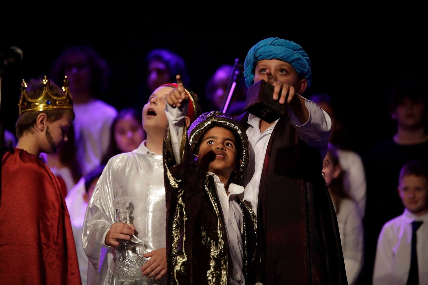 Children in nativity costumes perform on stage, including kings and wise men in colorful robes during a Christmas play.