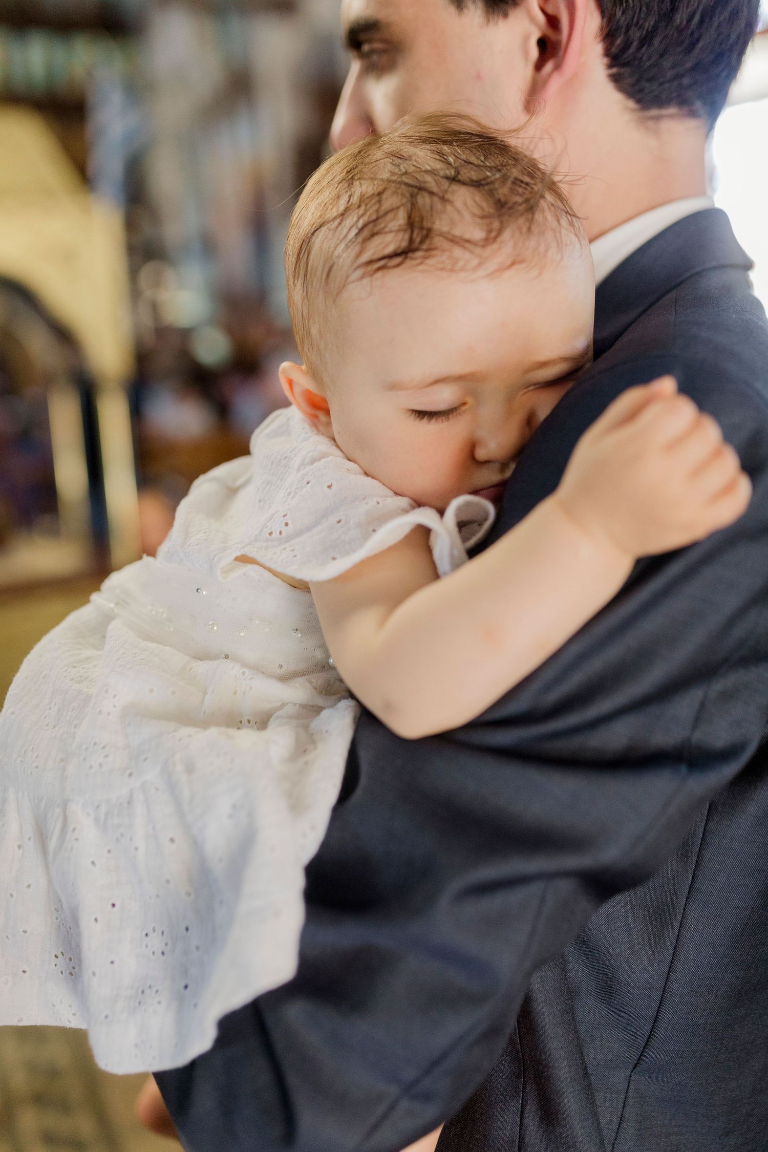 A baby in a white dress rests on a man's shoulder in a church during a baptism ceremony.