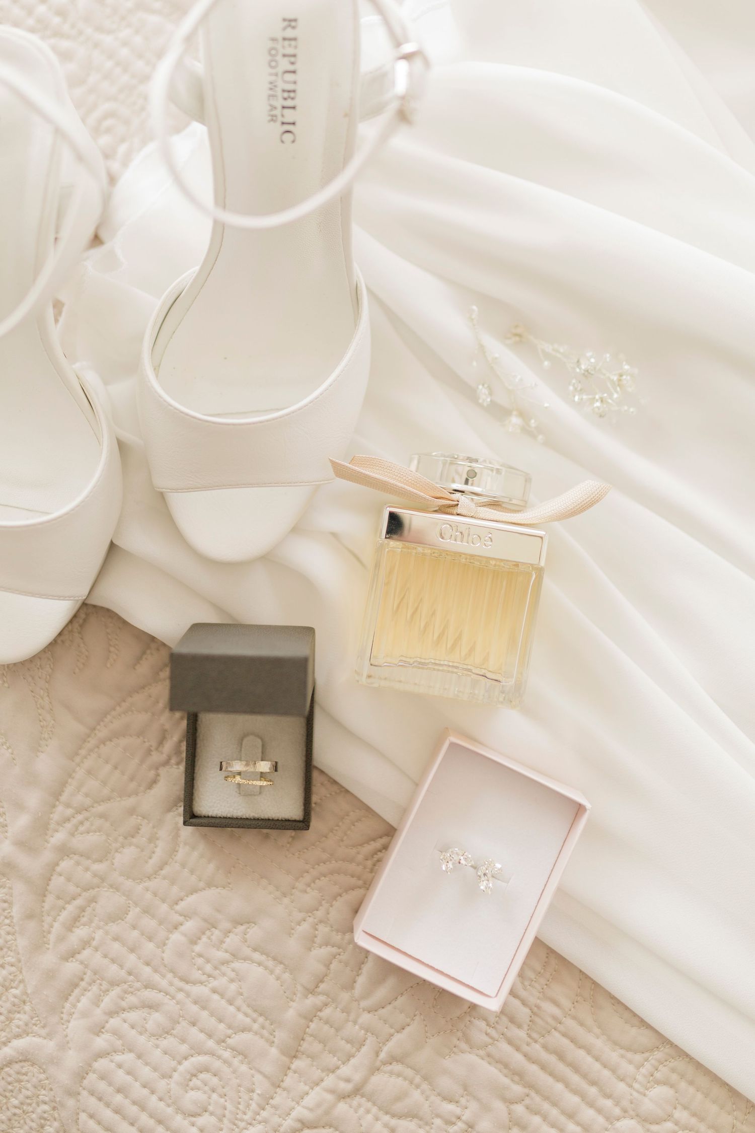 Elegant bridal flat lay with white heels, Chloe perfume, diamond rings in boxes, and flowing white wedding dress.
