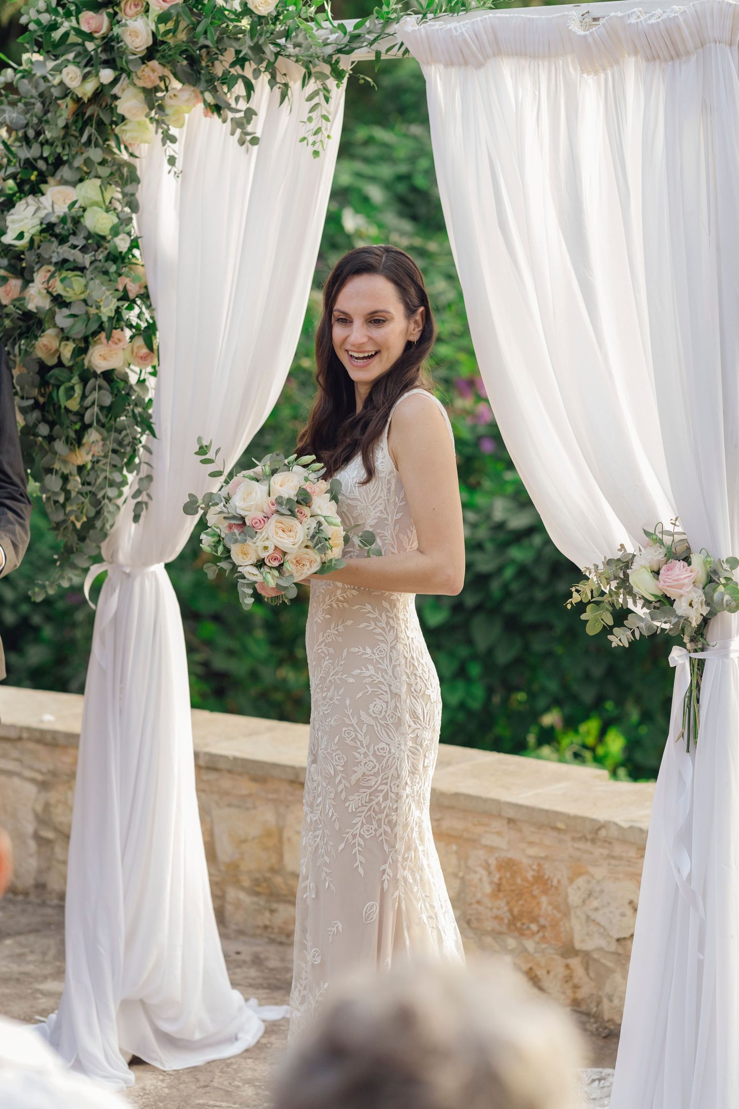 Smiling bride in beige lace gown holds bouquet under floral arch with white draping at outdoor wedding ceremony.