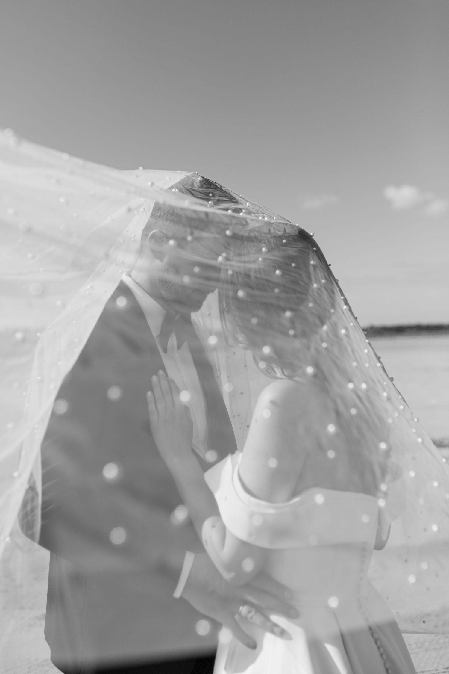 Black and white photo of bride and groom kissing under a polka dot veil on a beach.