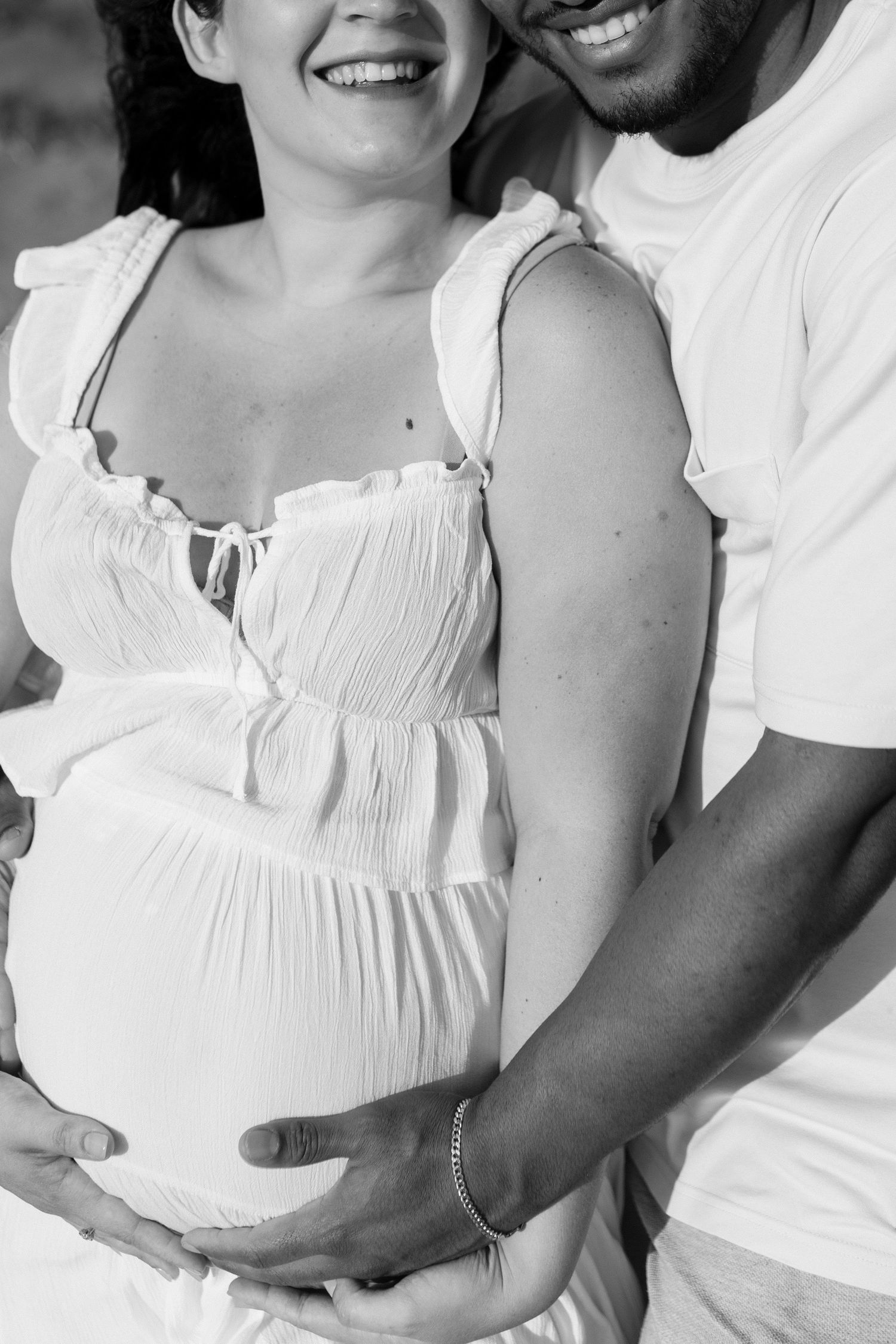 Black and white photo of a smiling pregnant couple tenderly cradling baby bump together.