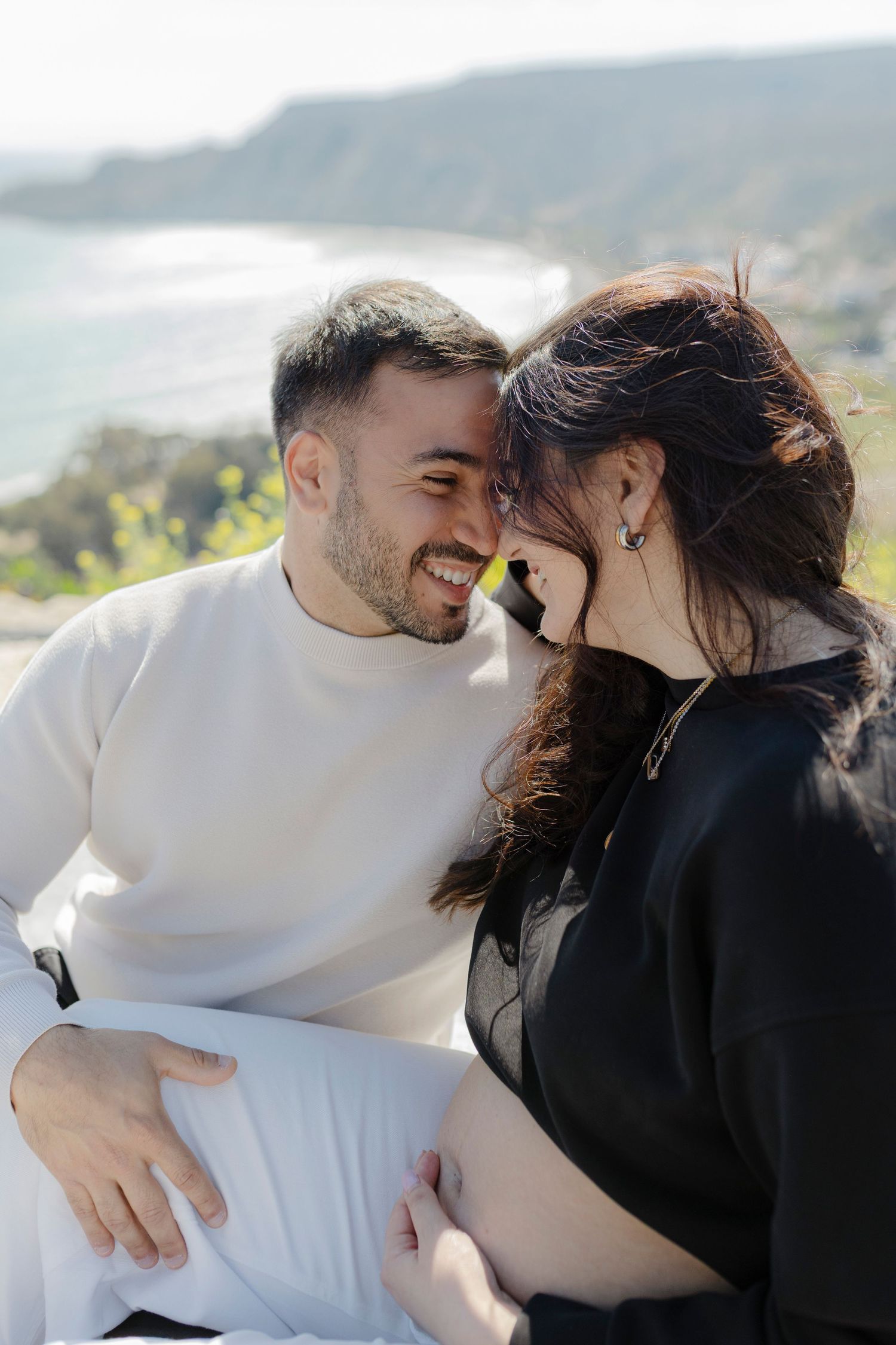 A smiling couple shares an intimate moment outdoors, with a coastal landscape in the background on a sunny day.