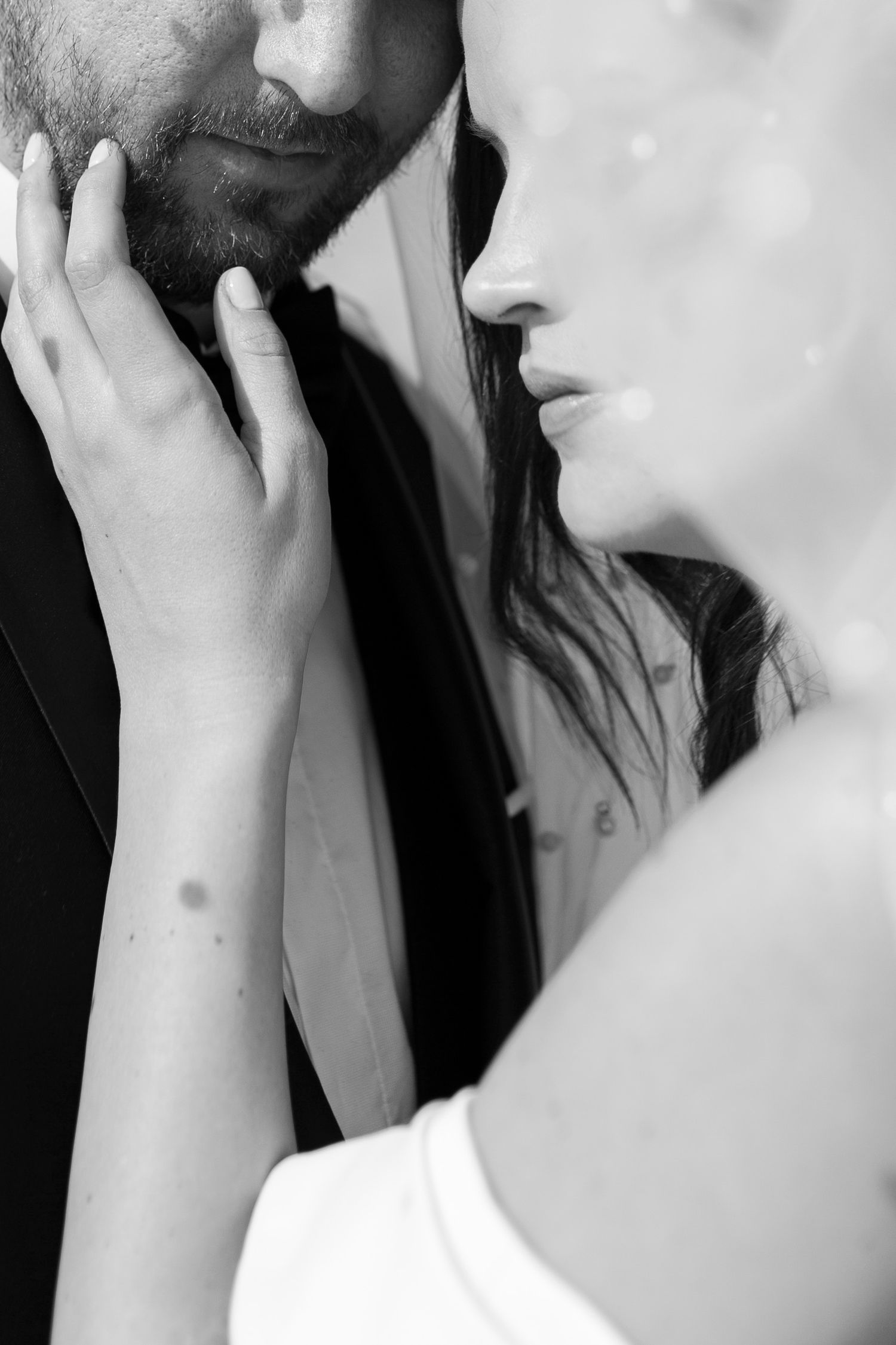 Intimate black and white close-up of bride gently touching grooms bearded face before a kiss.