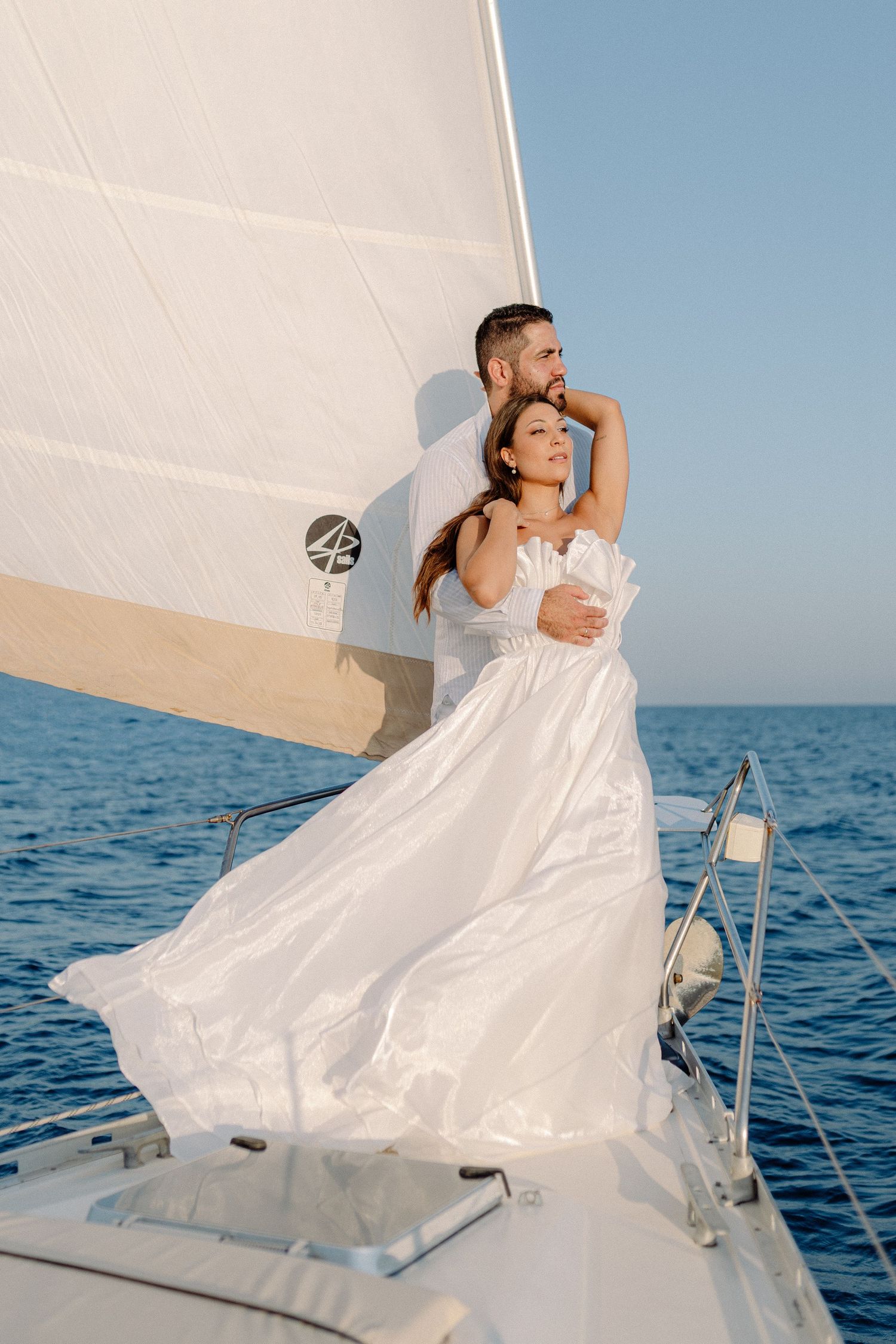 Bride in flowing white gown embraced by groom on sailboat with white sail and blue sea.
