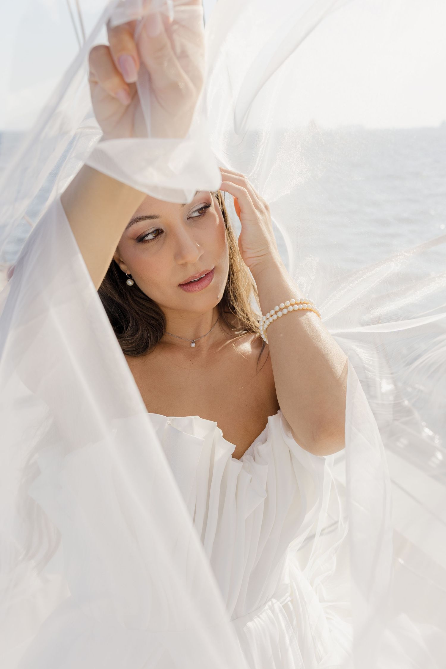 Elegant bride in white dress holding sheer veil with pearl bracelet and necklace near ocean.
