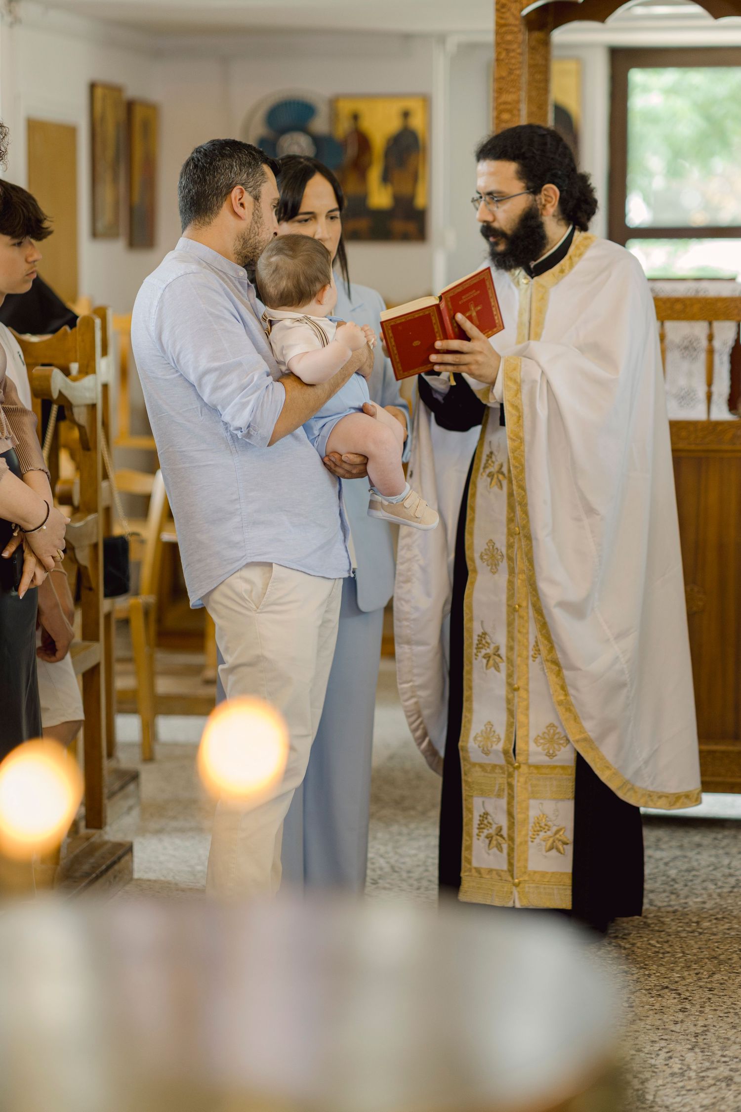 Orthodox priest reads from red book to couple holding baby during christening ceremony in bright church.