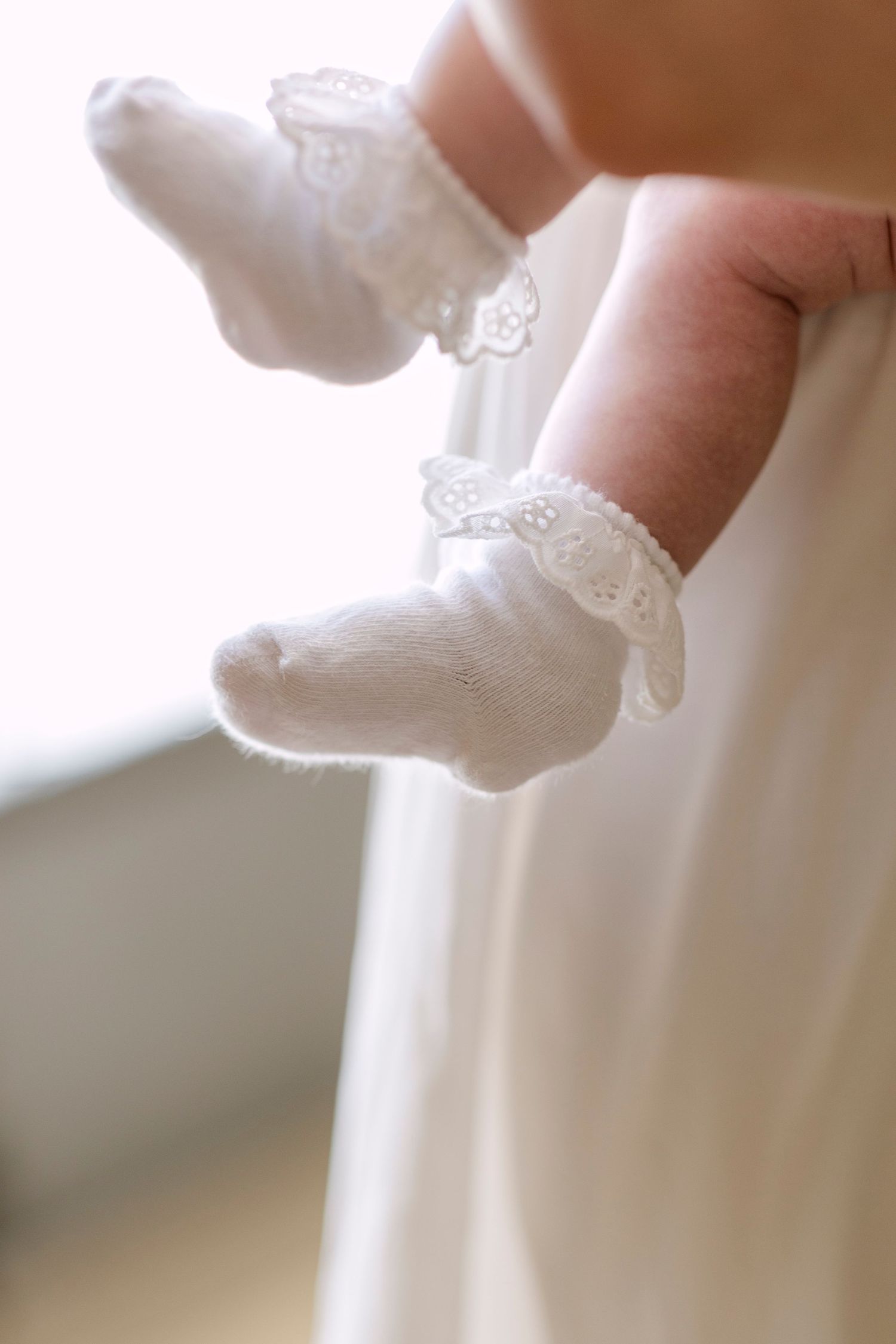 Close-up of baby feet wearing white lace-trimmed socks, held by an adult hand against a soft white background.