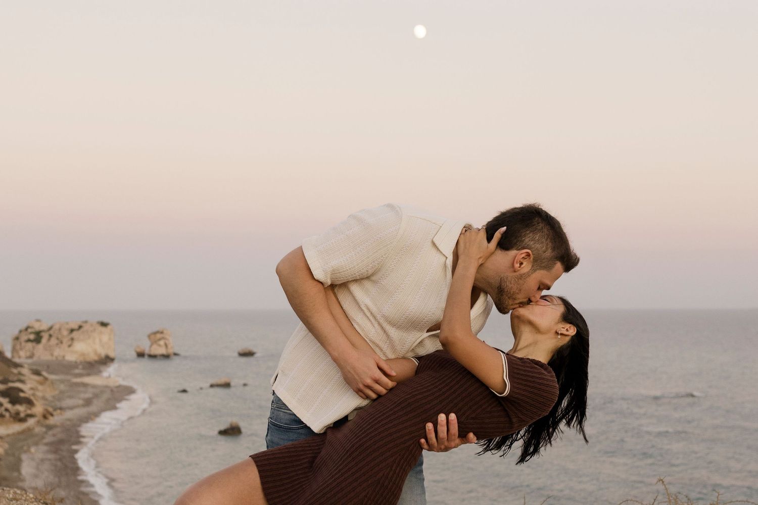 Romantic couple sharing a dip kiss on a rocky beach at dusk with a full moon in the pastel sky.