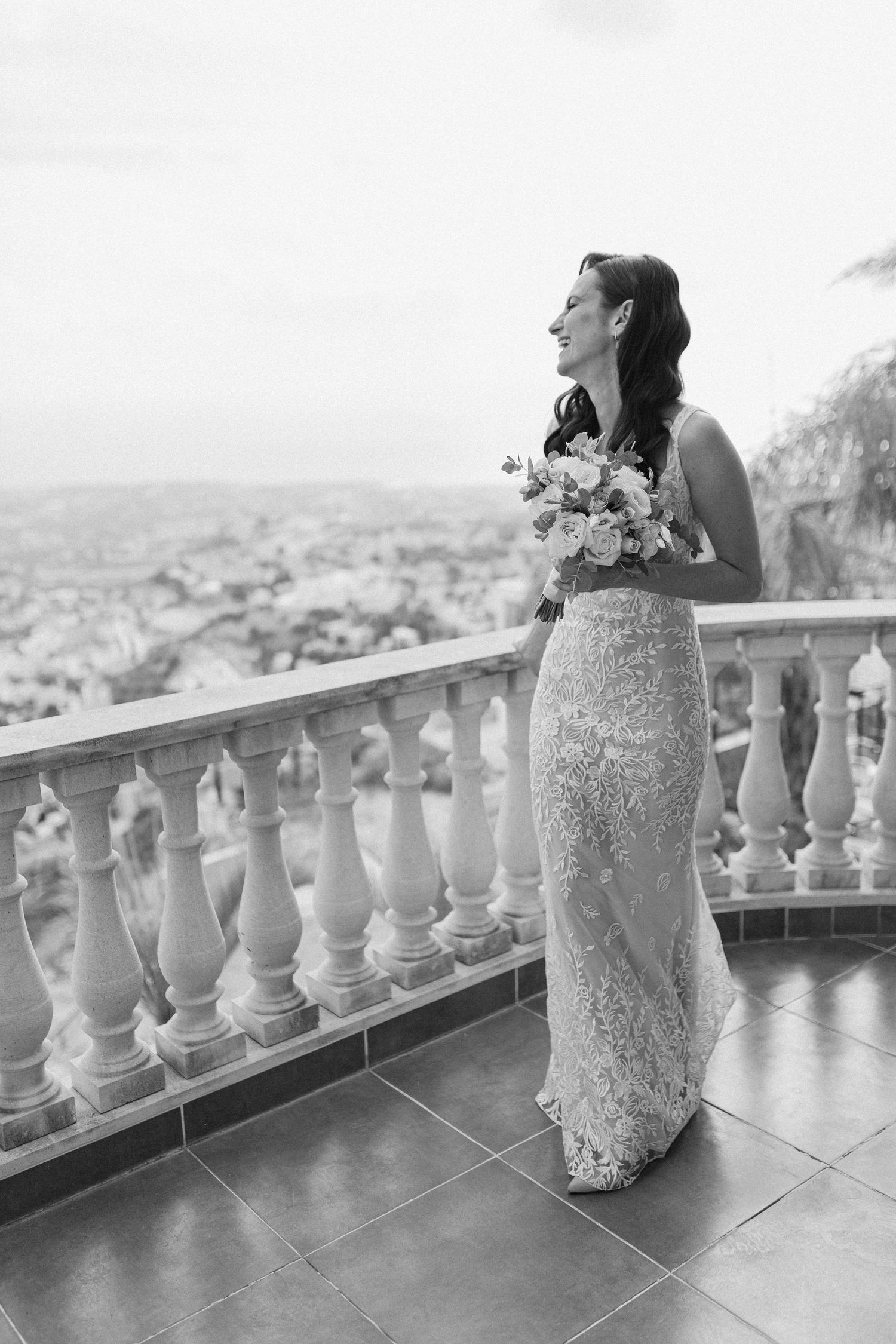 Black and white portrait of smiling bride in lace gown holding bouquet on scenic terrace with city views.