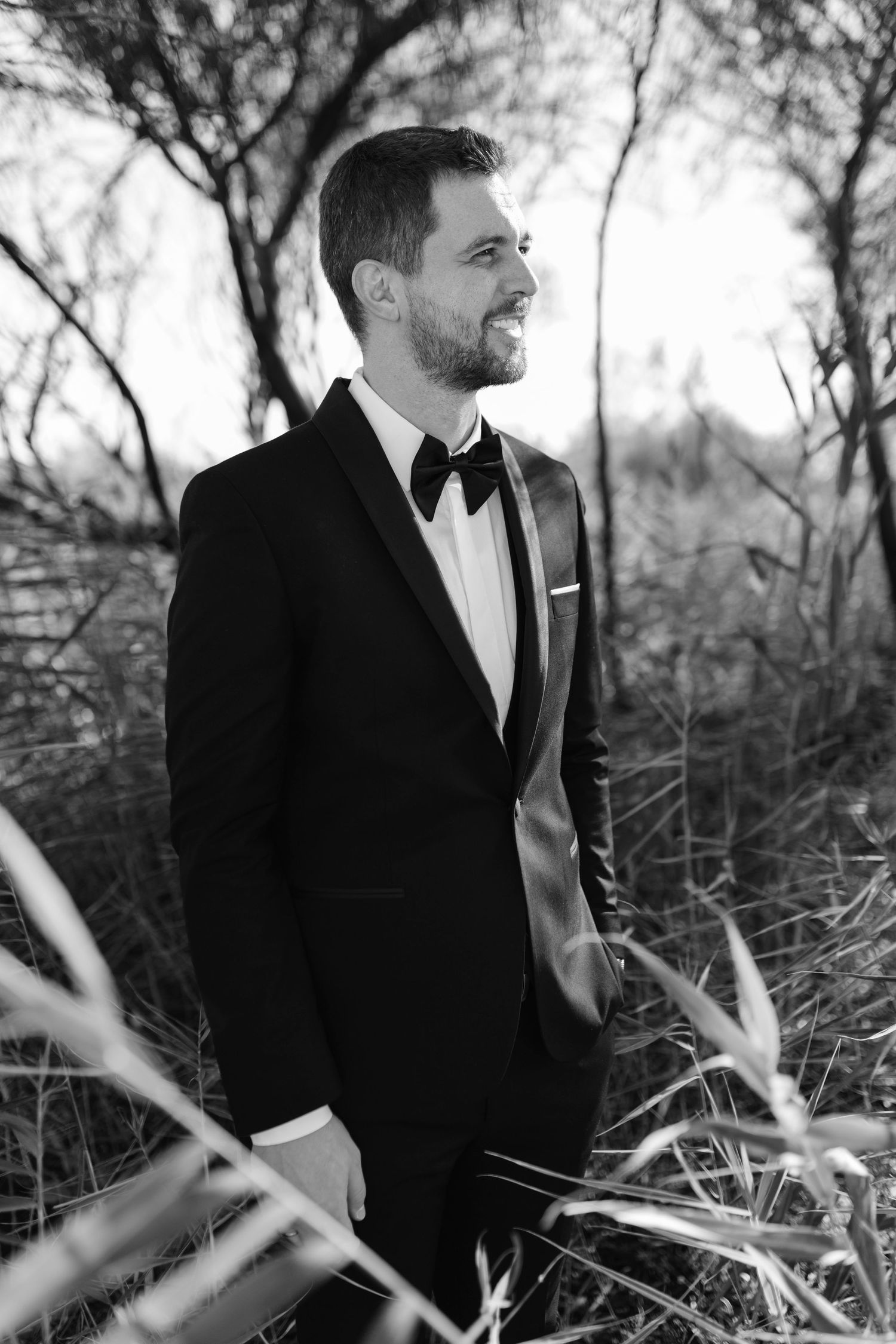 Black and white portrait of smiling groom in black tuxedo standing among tall grass and trees.