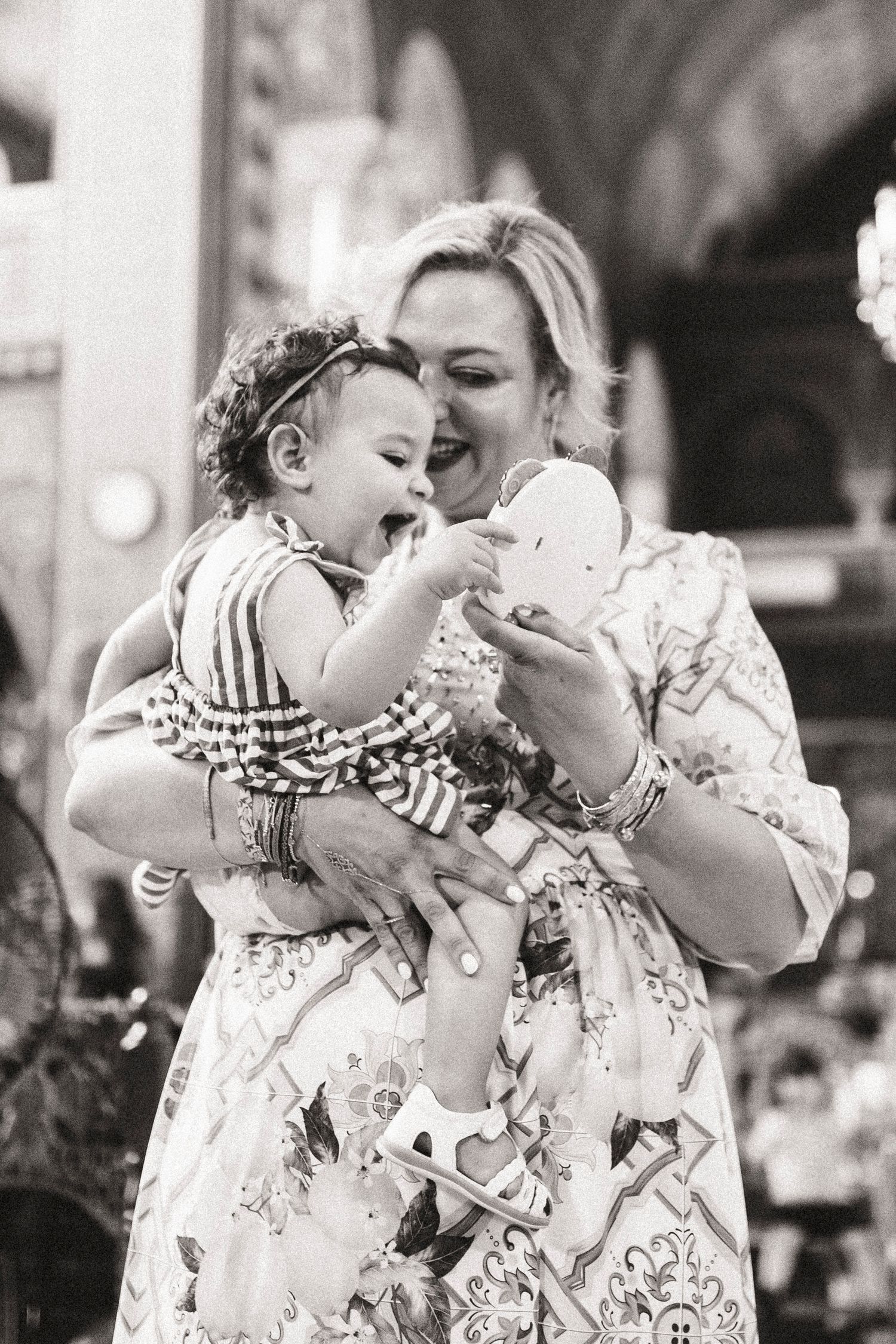 Black and white photo of a smiling woman holding a laughing baby girl in a striped dress at a celebration.