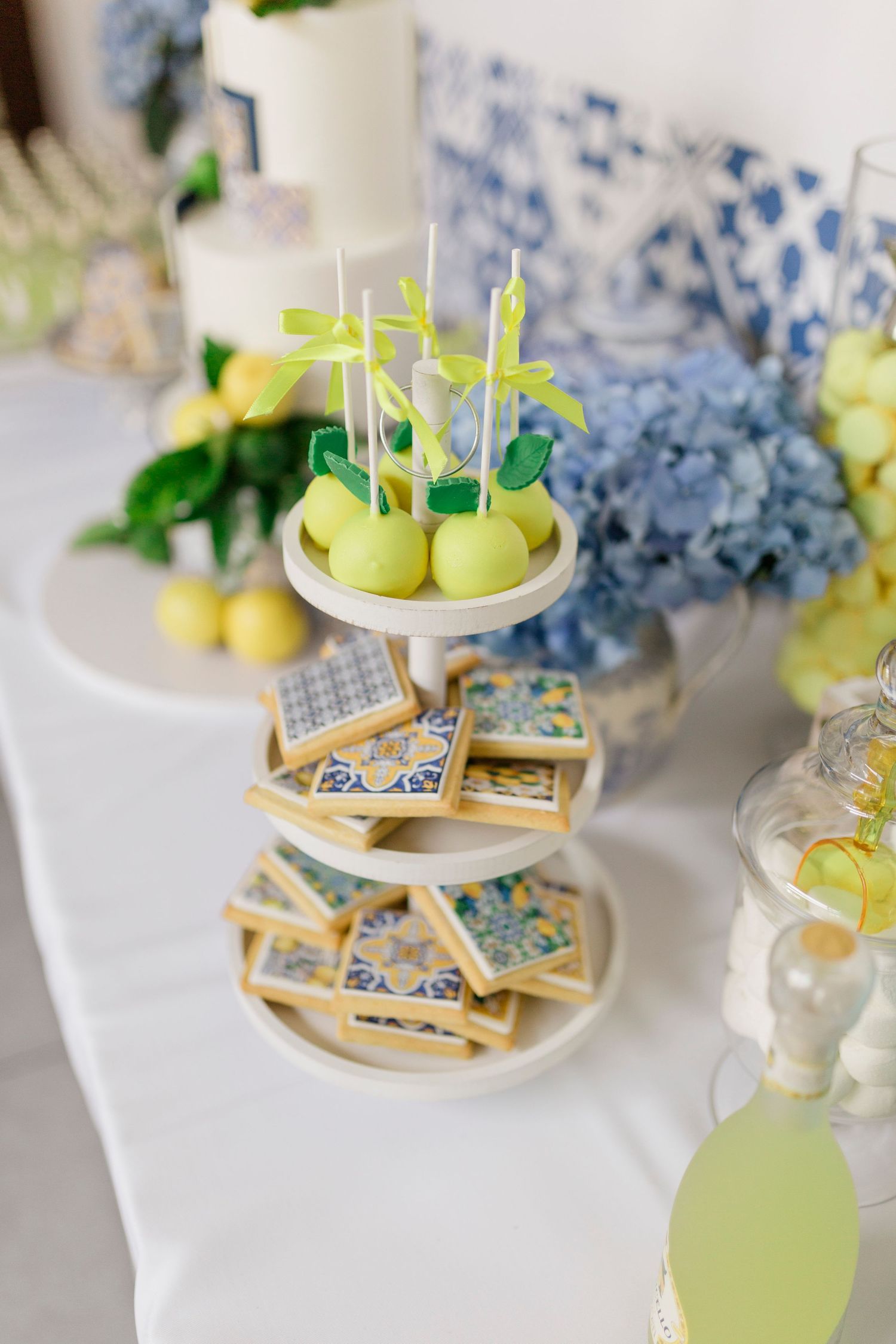 Elegant tiered stand with lemon cake pops and blue-yellow tile cookies on a white table with hydrangeas.