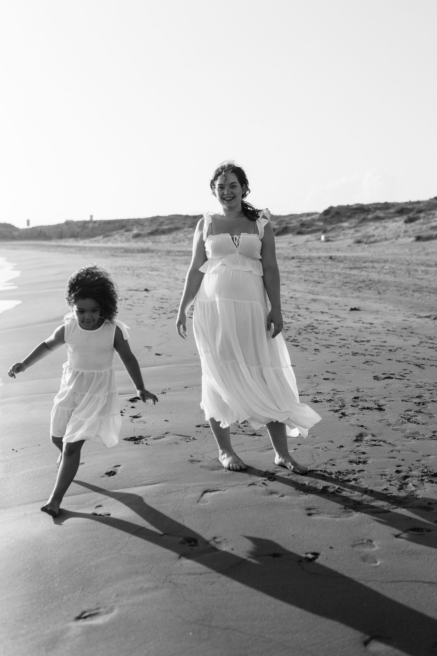 A mother and young daughter in white dresses walk barefoot on a sandy beach in a black and white photo.