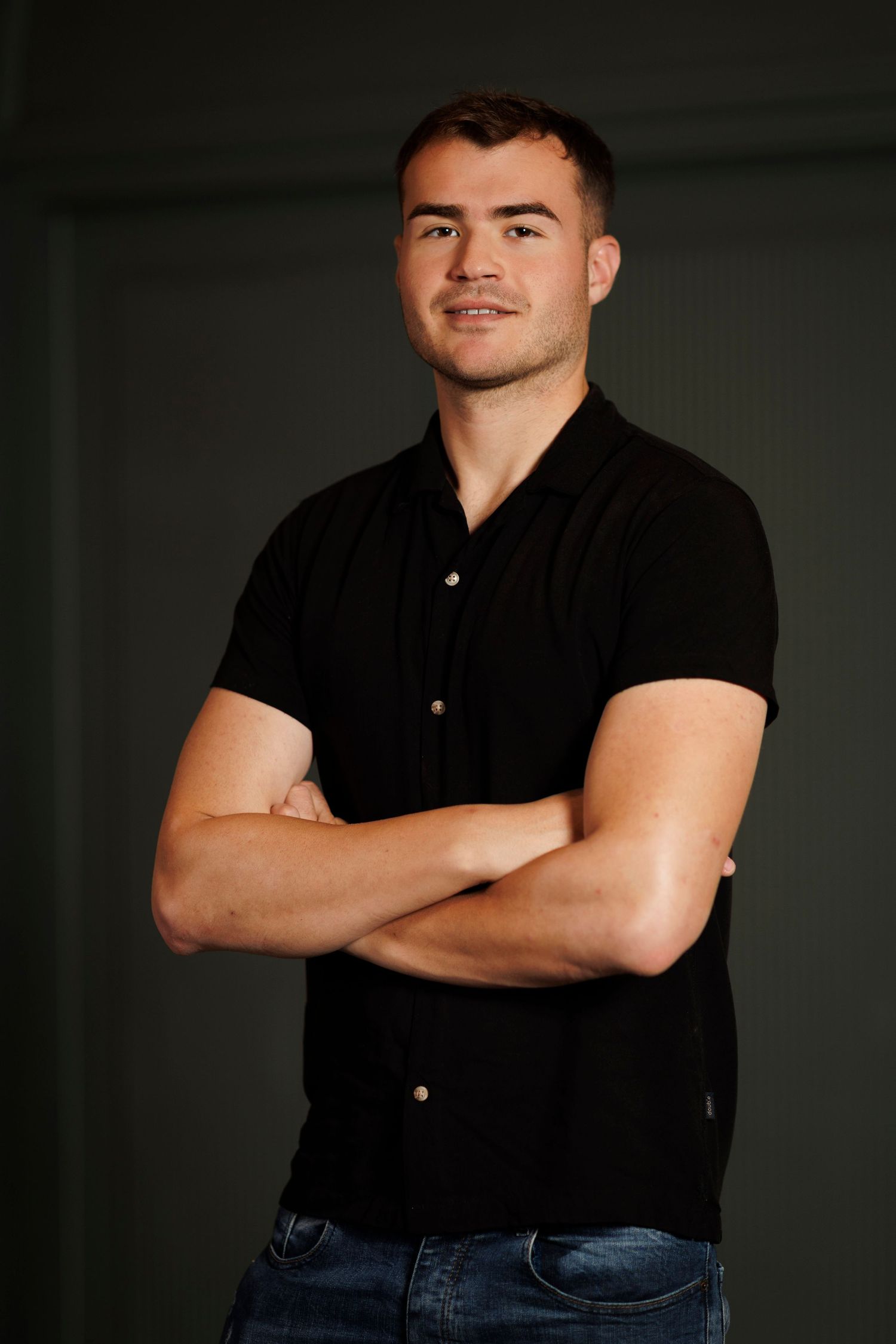 Young man in black polo shirt with arms crossed, smiling confidently against dark background.