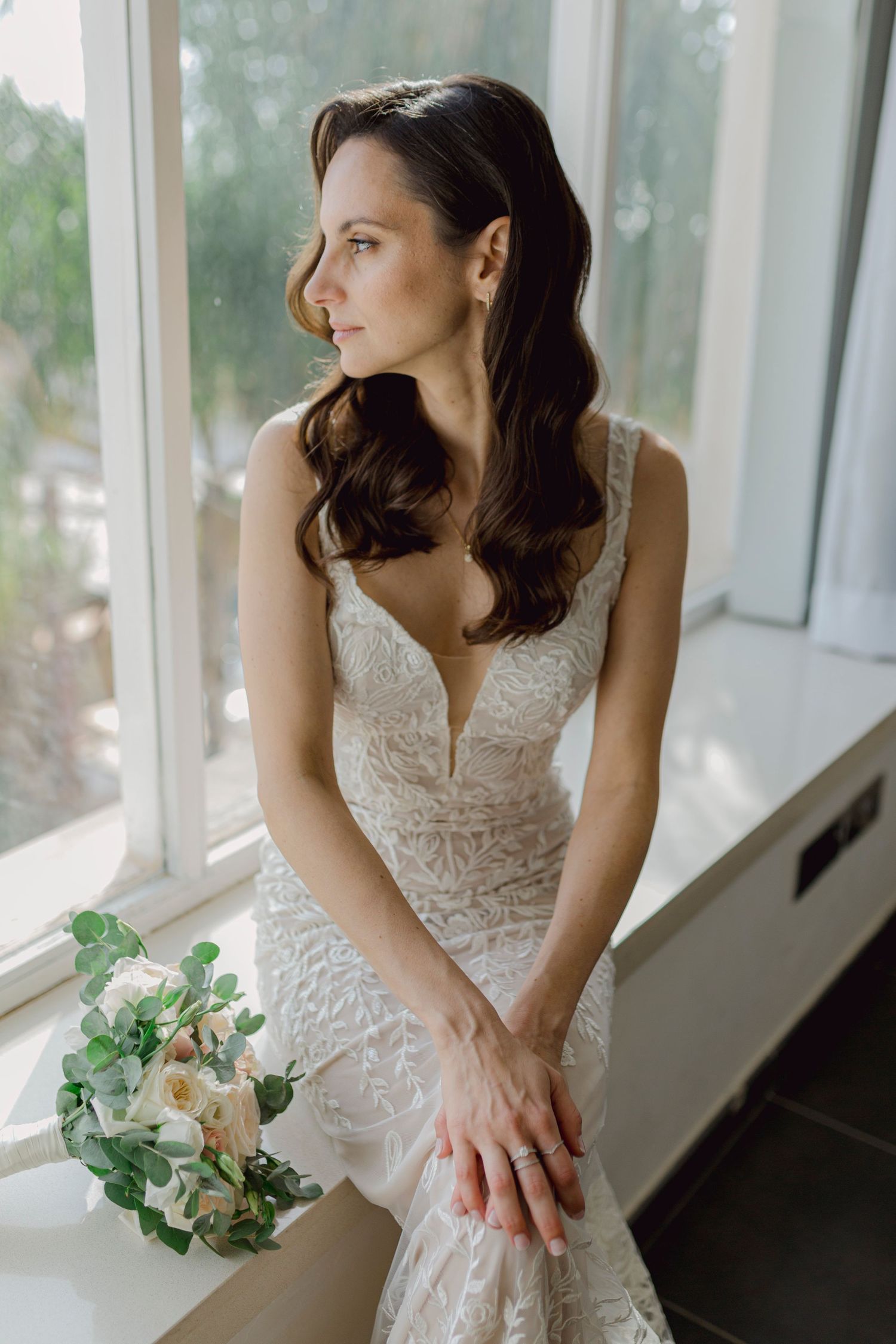 Bride in lace mermaid wedding gown holding white floral bouquet, seated by window in soft natural light.